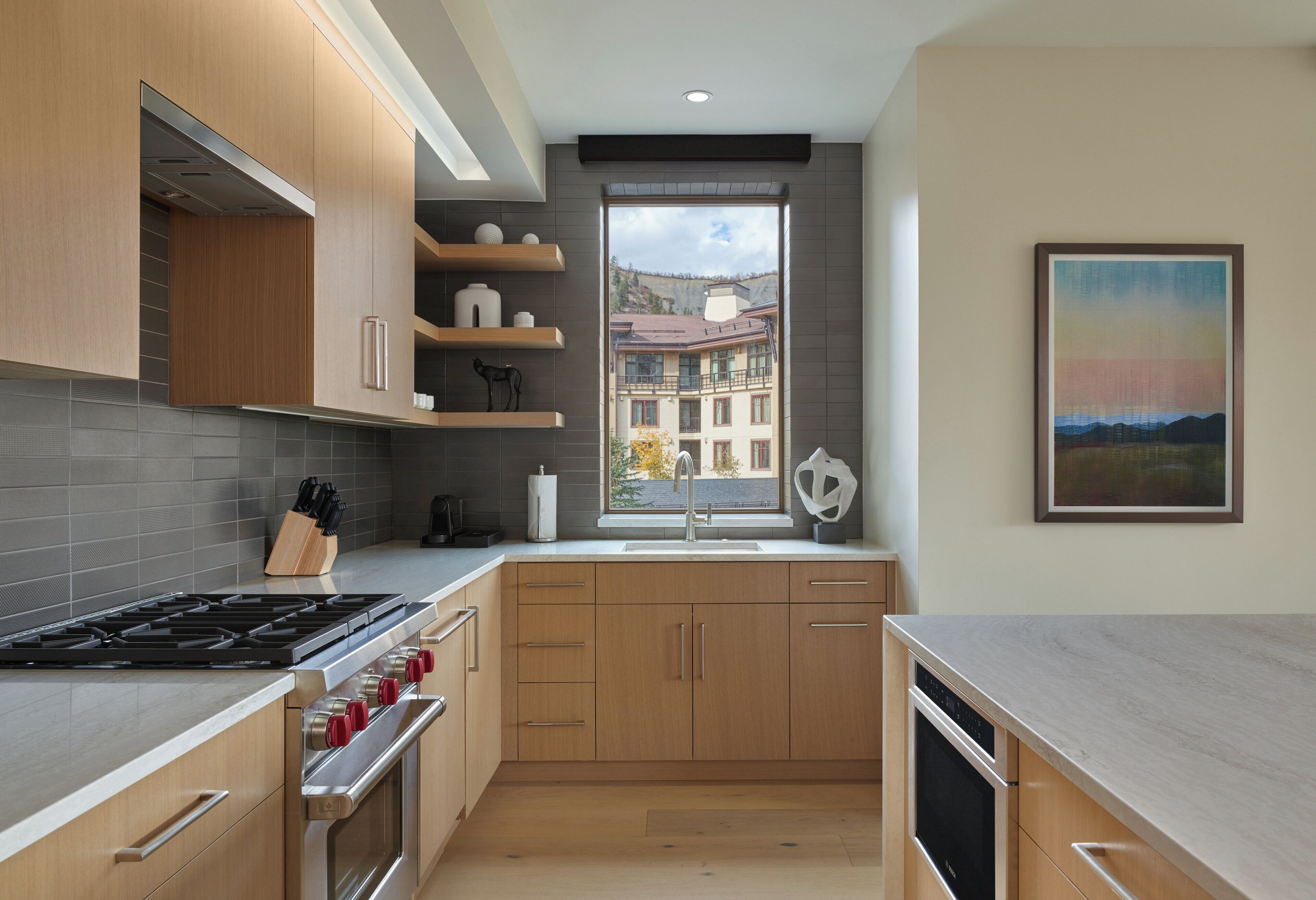 A modern kitchen with wooden cabinets, a stainless steel stove, and a window with a view of buildings.