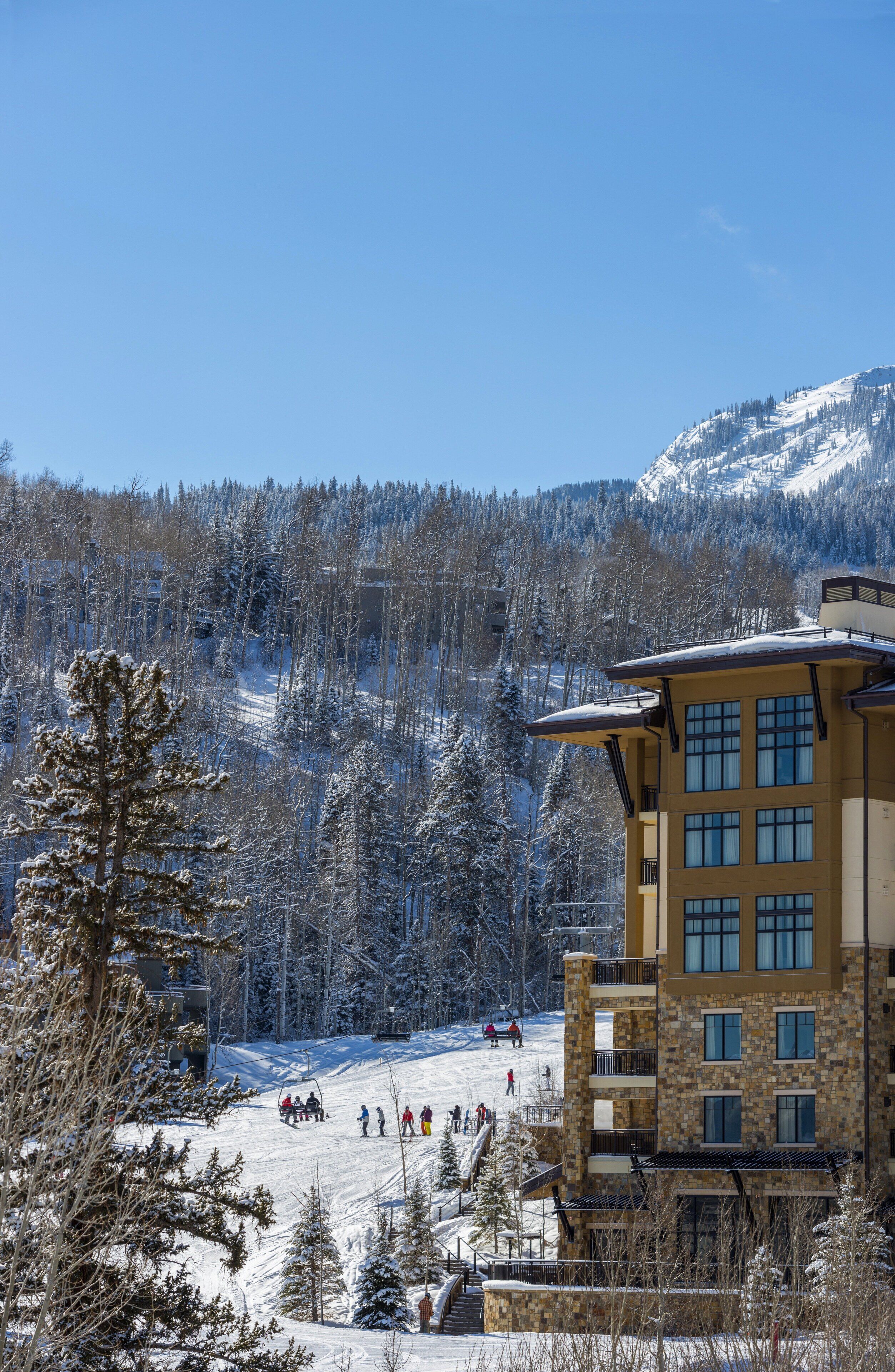 Un edificio de varios pisos con fachada de piedra y ventanas grandes, rodeado de árboles cubiertos de nieve y una pista de esquí.