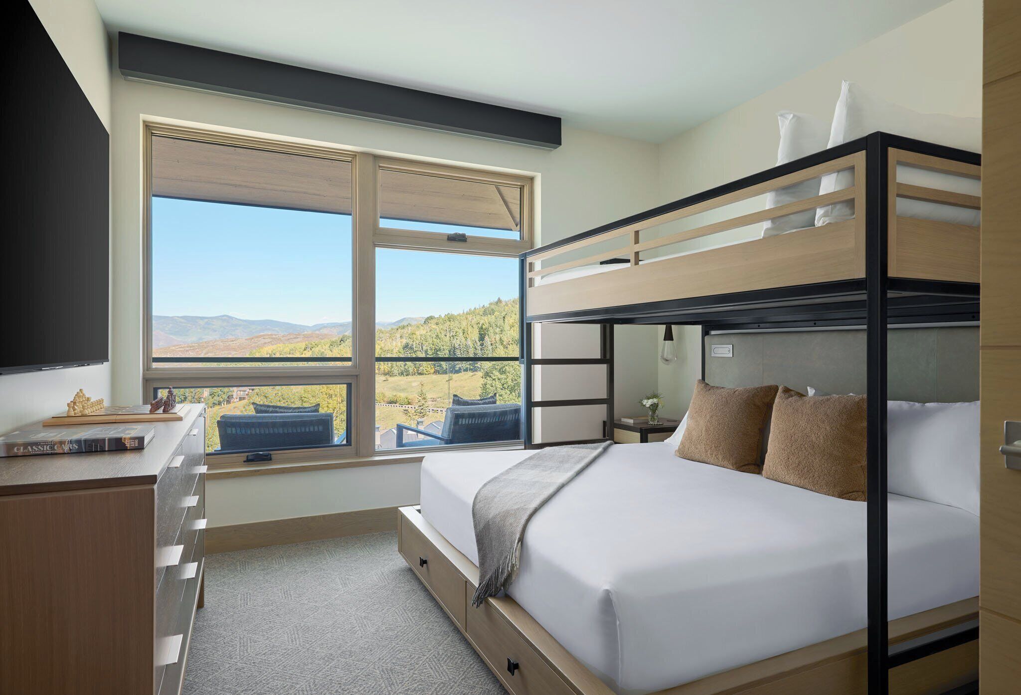 A bunk bed room with a desk, a window with a view of the mountains, and a small table with books.