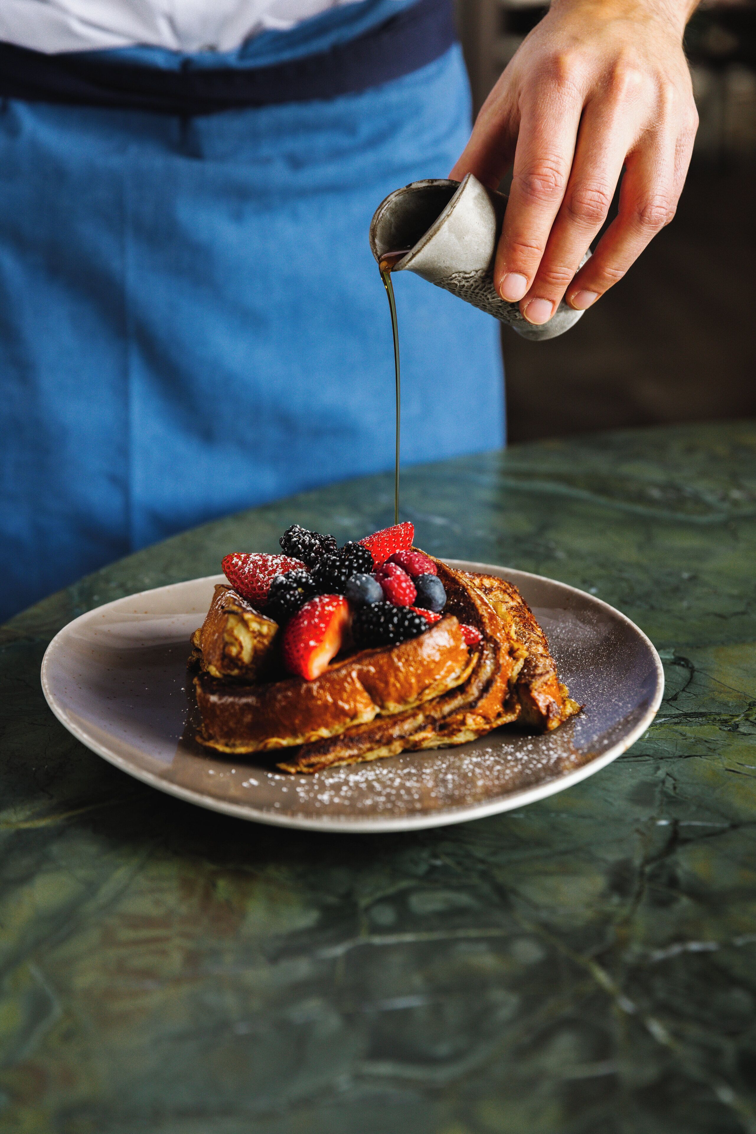 A person pouring syrup over a breakfast dish with strawberries and blueberries.