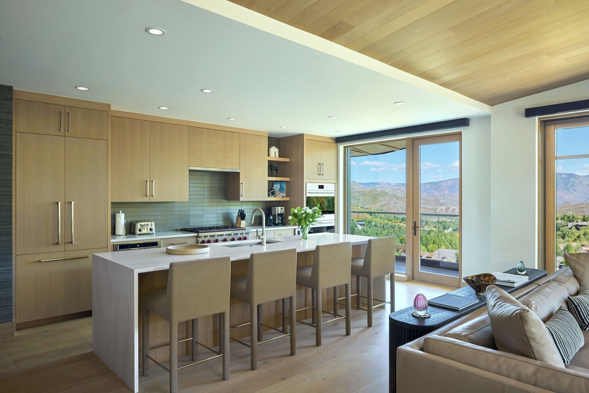A modern kitchen with a central island, bar stools, and a view of mountains through large windows.