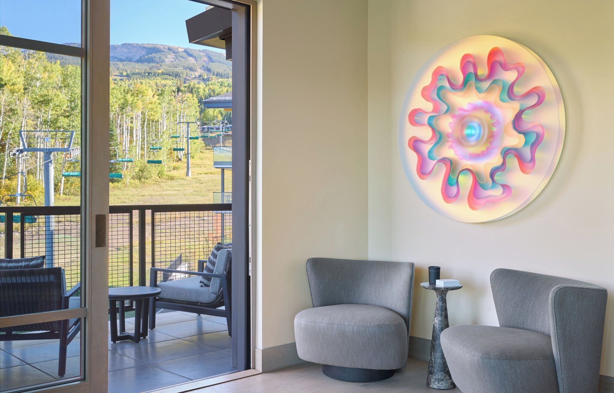 A room with two grey armchairs, a small round table with a vase, and a colorful wall art piece.