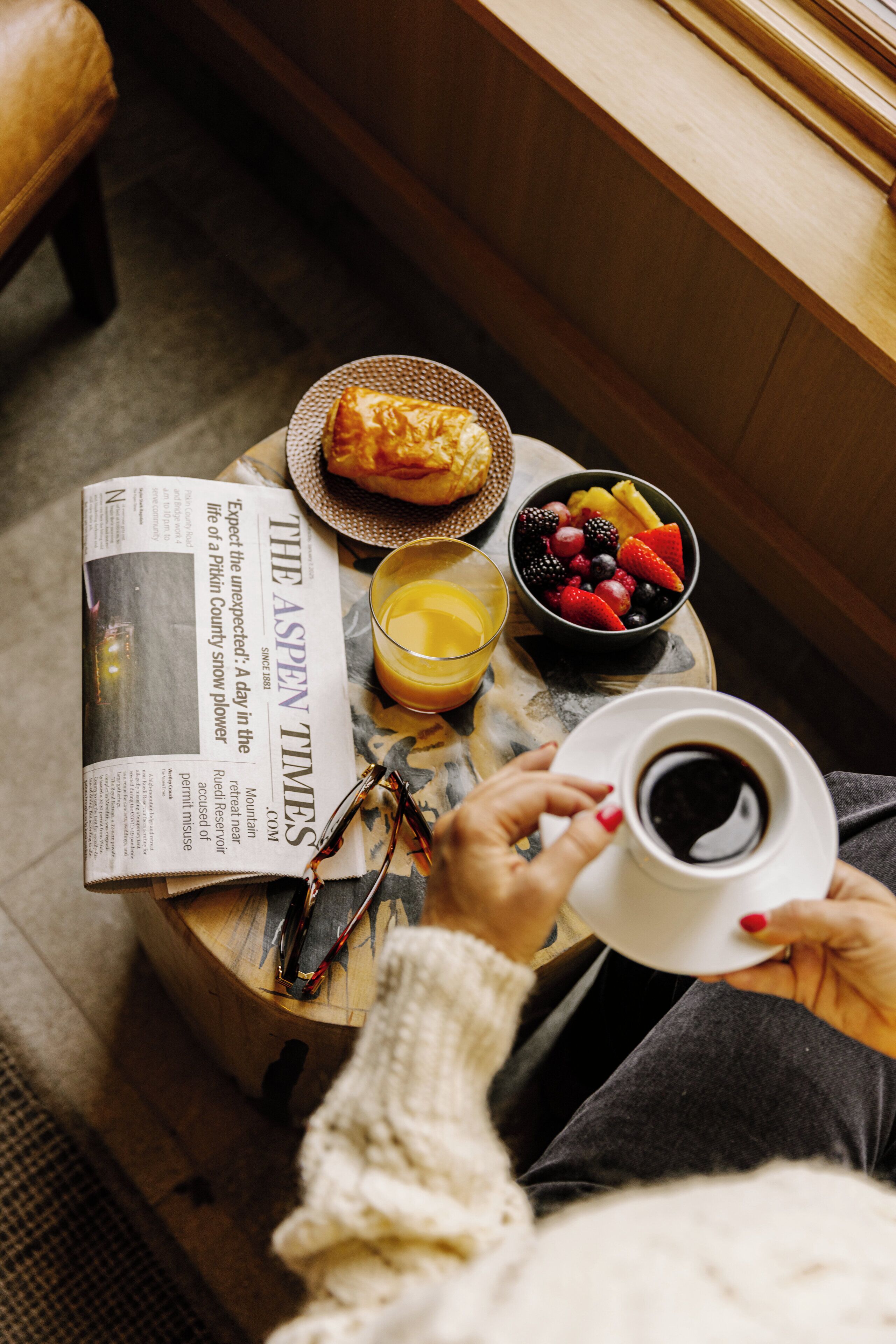 Eine Person hält eine Kaffeetasse, eine Zeitung und einen Obstkorb auf einem runden Holztisch.