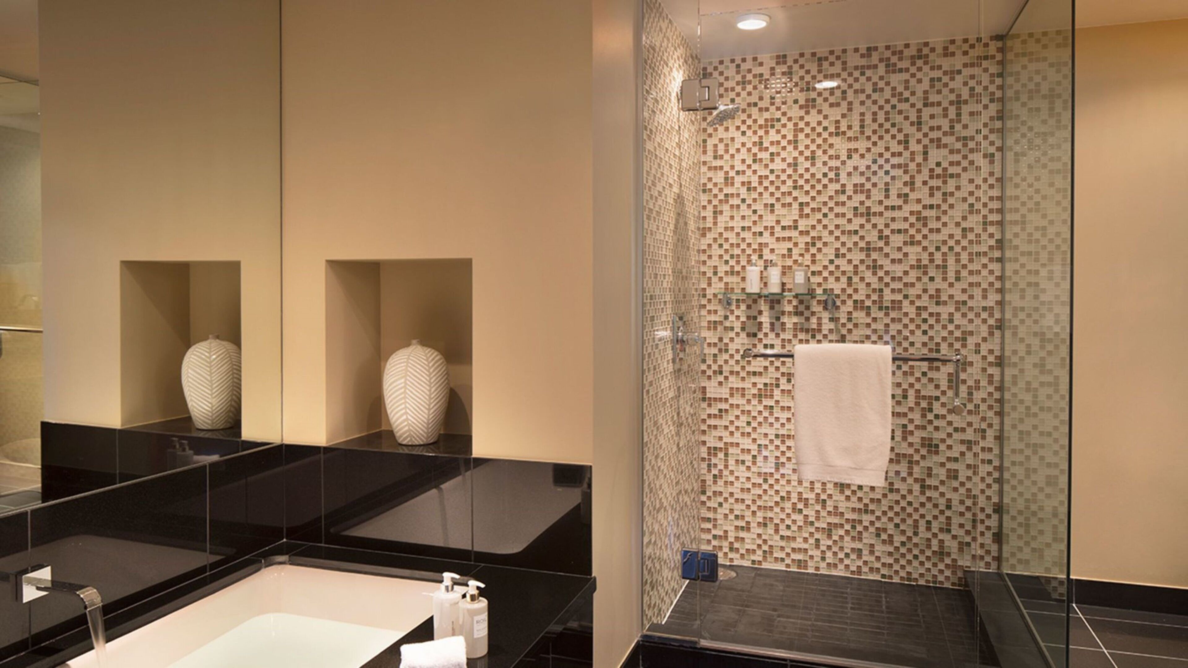 A modern bathroom with a large mirror, two vases, a bathtub, and a shower area with mosaic tiles.