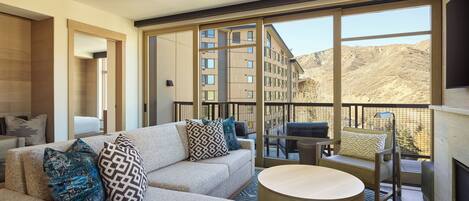 A modern living room with a sofa, coffee table, and chairs, large windows offering a view of a building and hills.