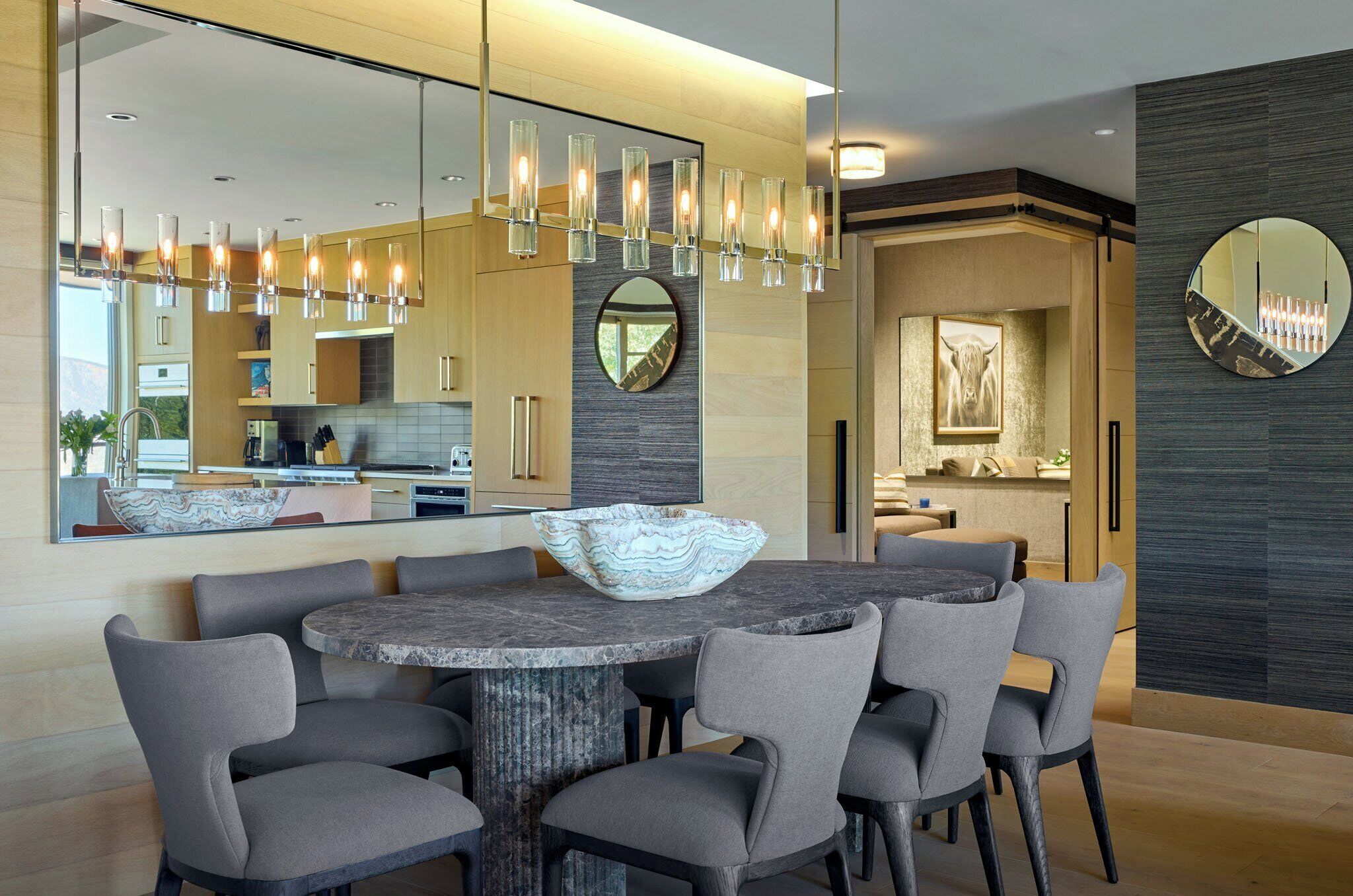 A modern dining area with a round table, grey upholstered chairs, and a decorative bowl centerpiece. A large mirror reflects the room, and a chandelier hangs above.