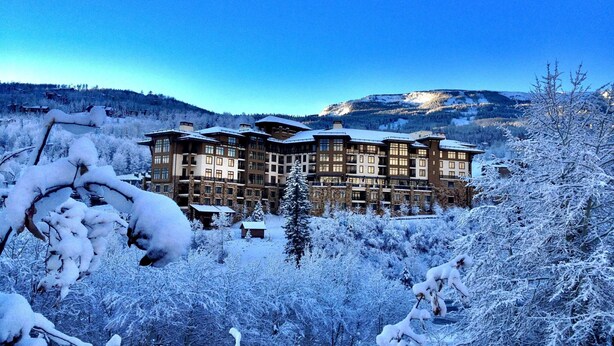 Um hotel de vários andares com fachada simétrica, rodeado por árvores cobertas de neve e um cenário montanhoso.