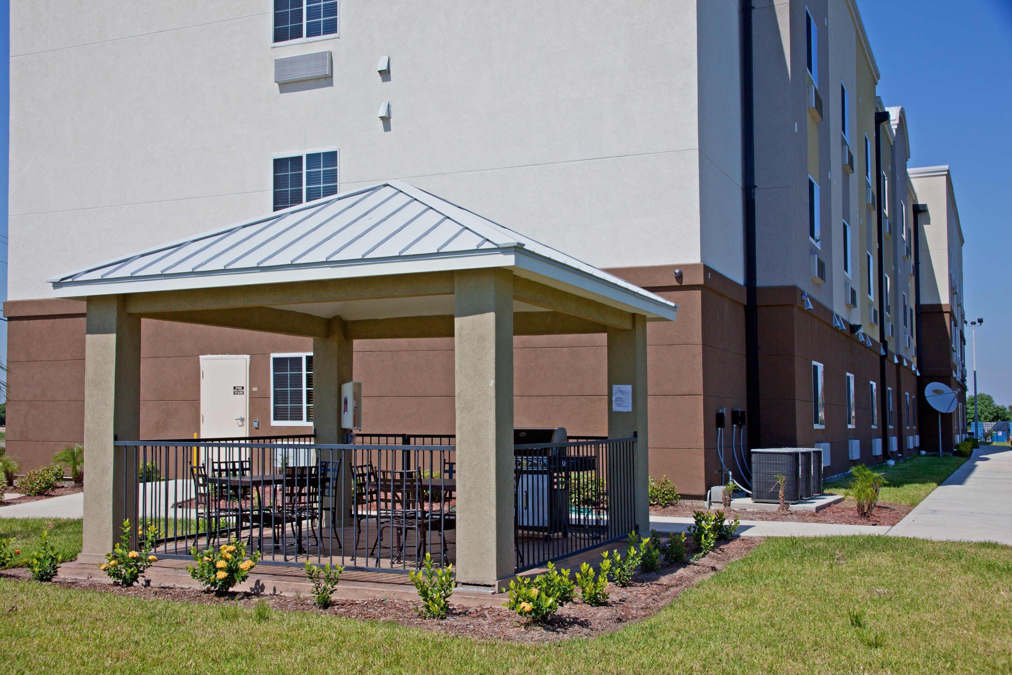 A covered outdoor seating area with tables and chairs, a building entrance, and a small garden with plants.