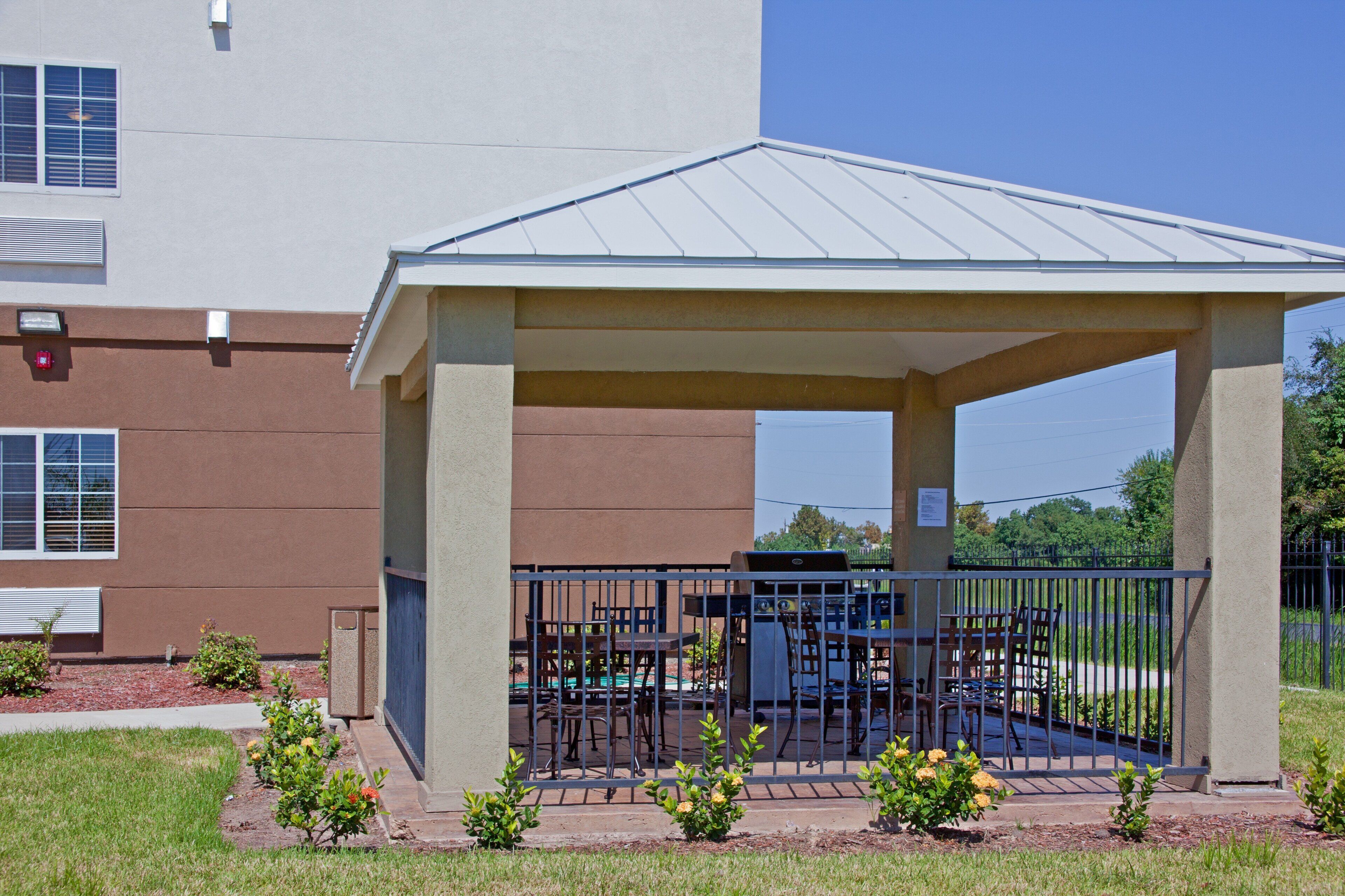 A covered outdoor seating area with a grill, surrounded by a metal fence, in front of a building with a brown facade and a flat roof.
