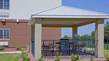 A covered outdoor seating area with a grill, surrounded by a metal fence, in front of a building with a brown facade and a flat roof.