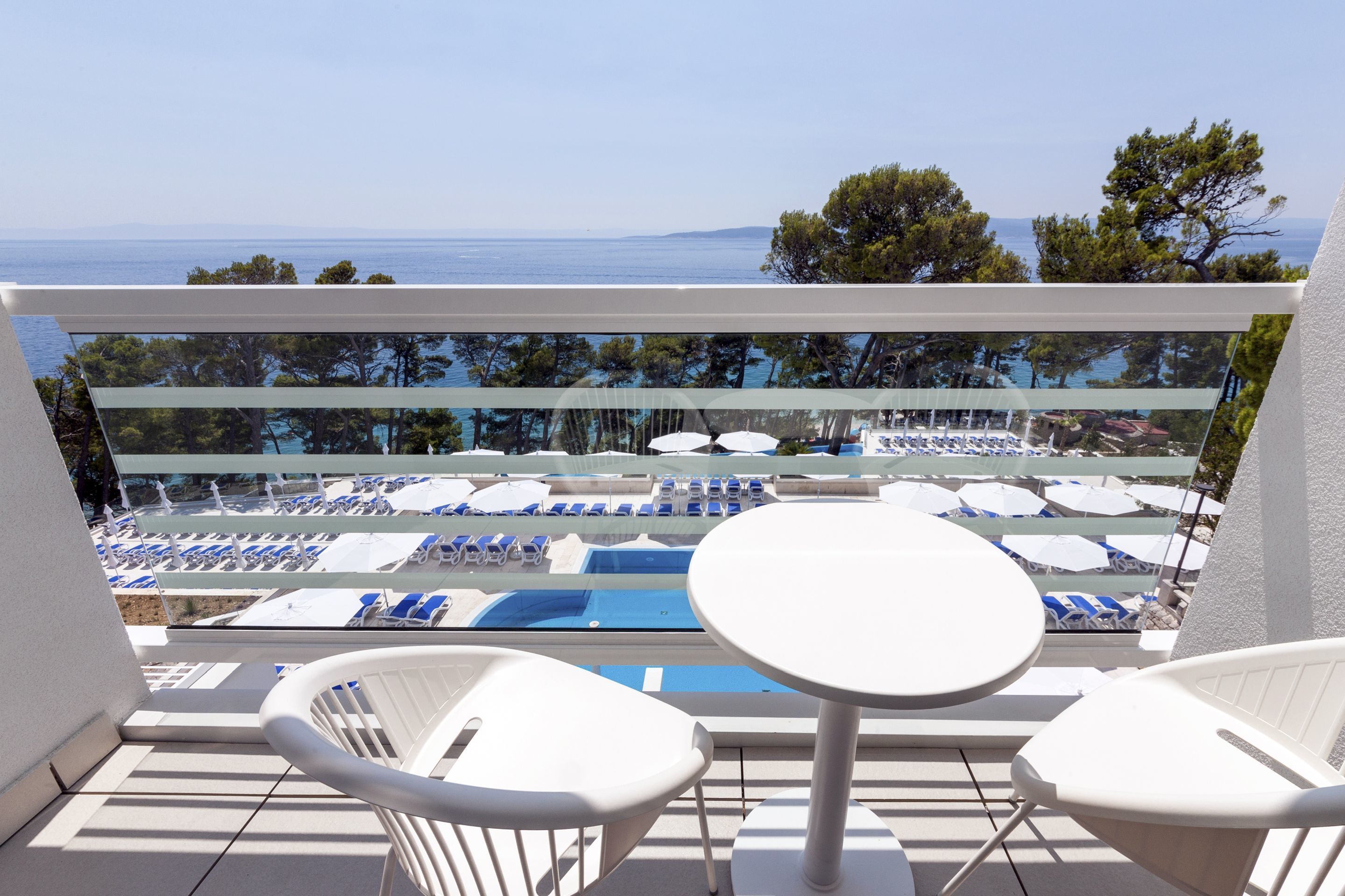 A balcony with white chairs and a table, overlooking a pool area with blue lounge chairs and umbrellas.