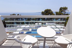 Ein Balkon mit weißen Stühlen und einem Tisch, der auf einen Poolbereich mit blauen Liegestühlen und Sonnenschirmen blickt.