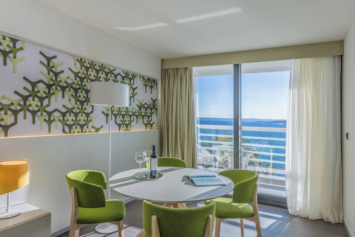 A modern room with a round table, green chairs, a wine bottle, and a view of the ocean.