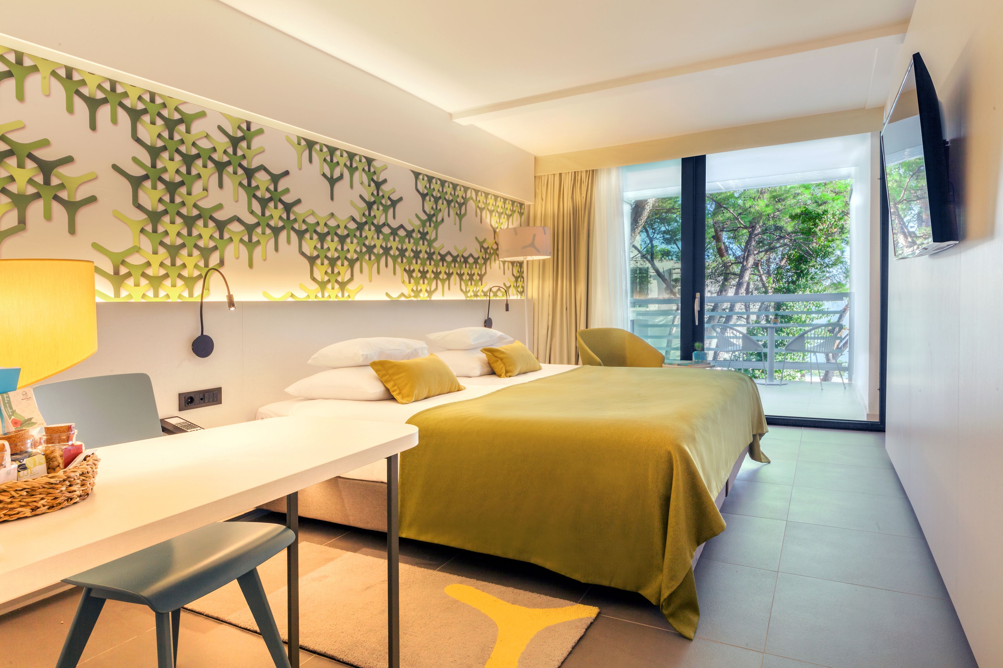 Ein modernes Hotelzimmer mit einem Bett, einem Schreibtisch mit einem Laptop, einem Stuhl und einem großen Fenster mit Blick auf Grünflächen.