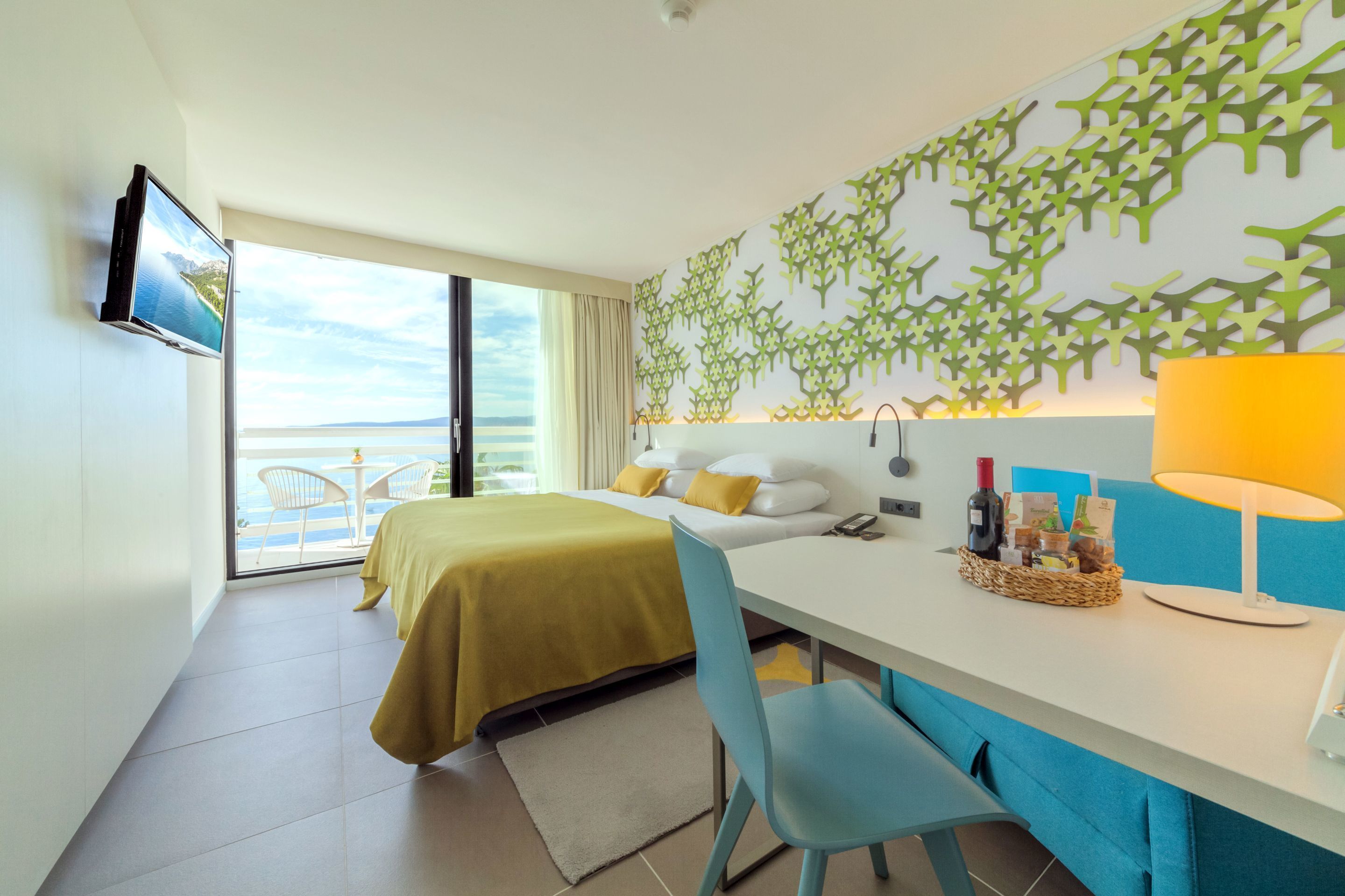 A hotel room with a bed, a desk with a lamp, a chair, a TV, and a view of the sea.