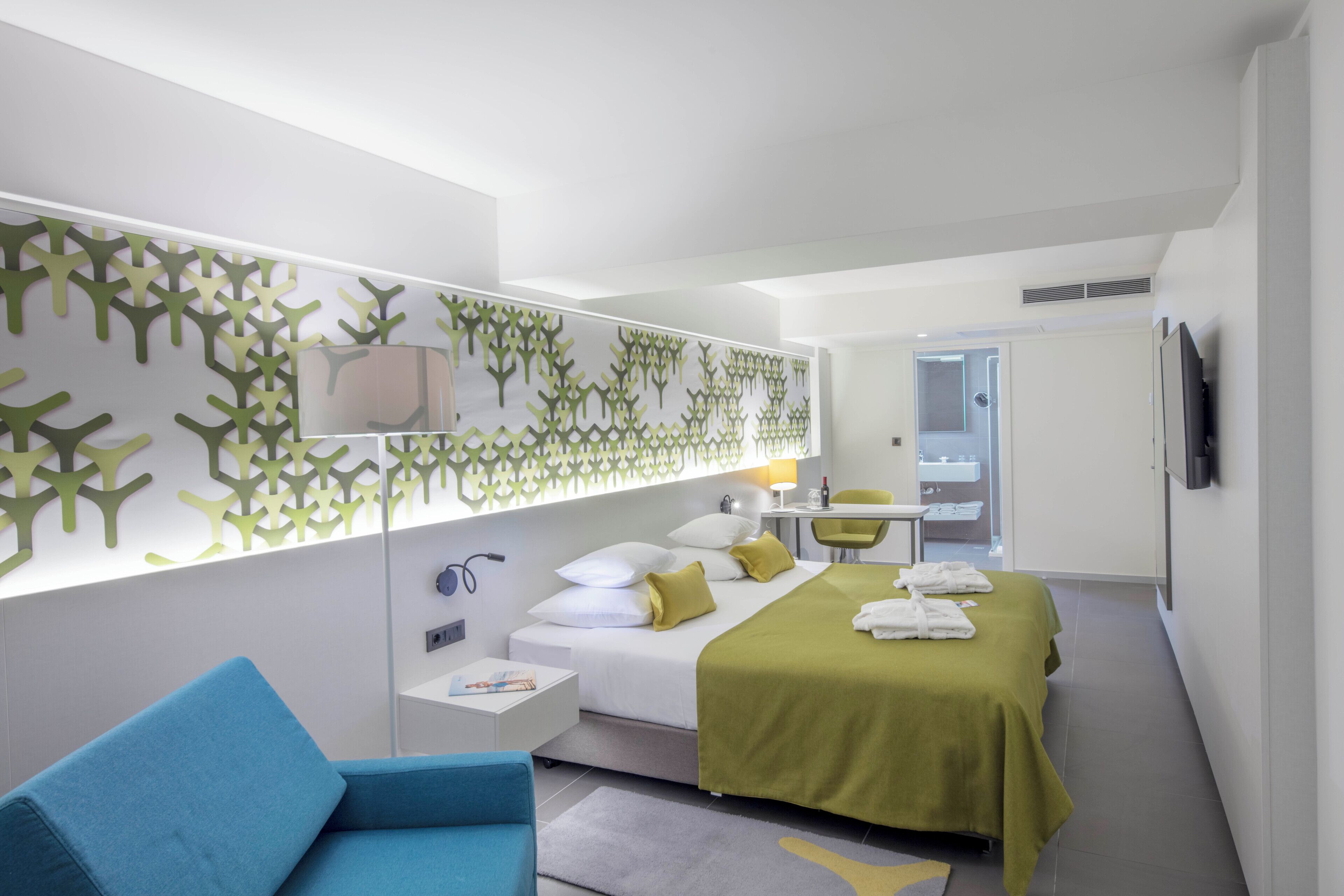 A modern hotel room with a bed, a desk, a chair, and a bathroom visible through an open door.