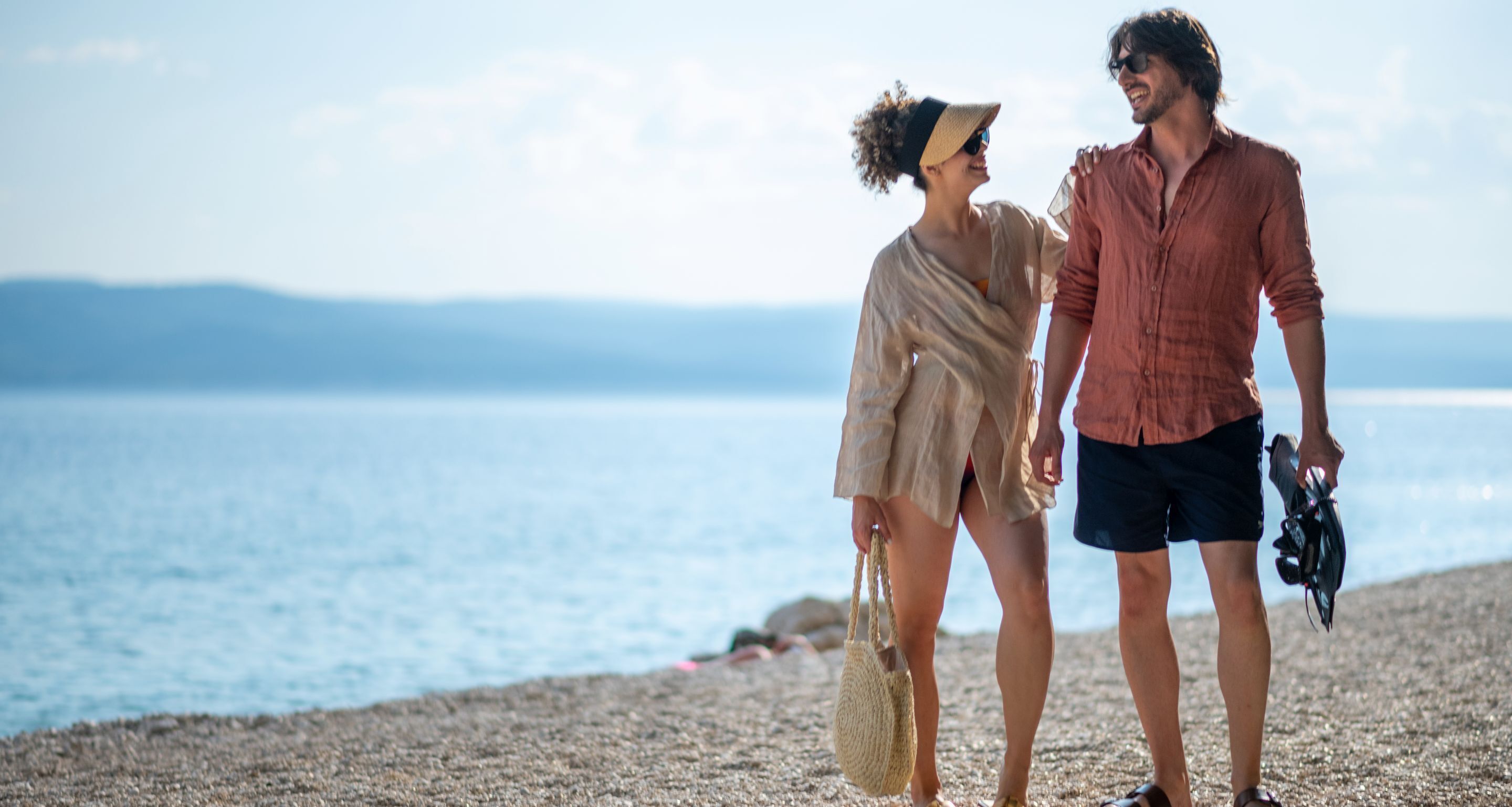A couple walking on a pebbled beach with a view of the sea and distant mountains.