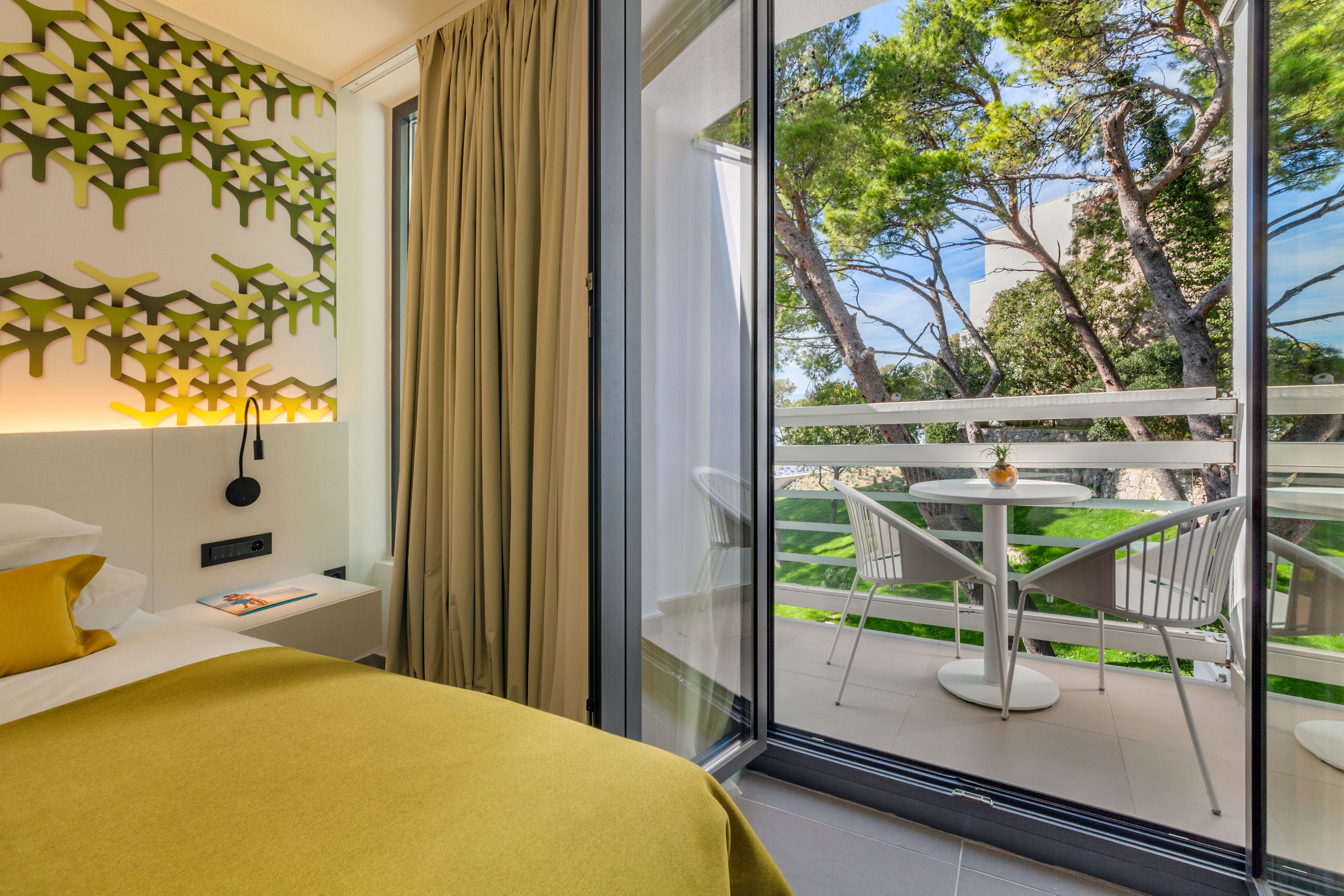 A hotel room with a bed, a nightstand, a wall-mounted lamp, a window with a view of a balcony, and a decorative wall panel.