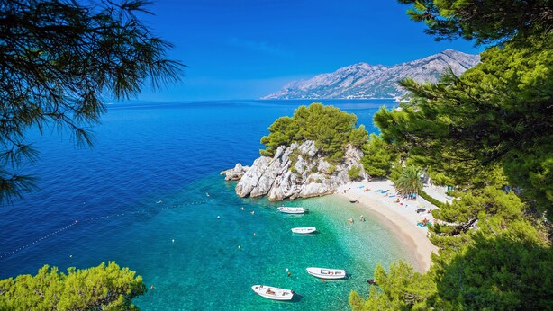 A coastal view with clear turquoise waters, a rocky outcrop with trees, and a beach with umbrellas and boats.
