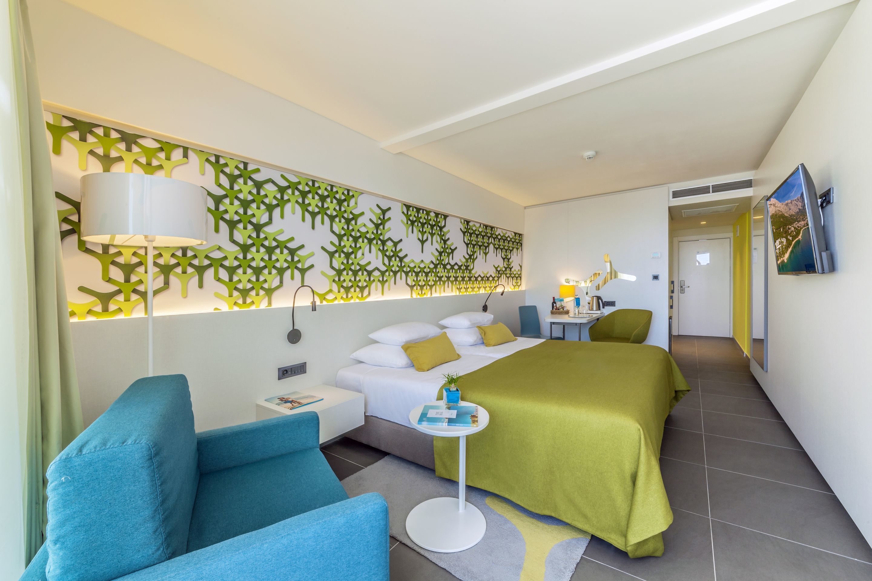 Een moderne hotelkamer met een bed, twee blauwe fauteuils, een klein tafeltje en een decoratief wandpaneel.