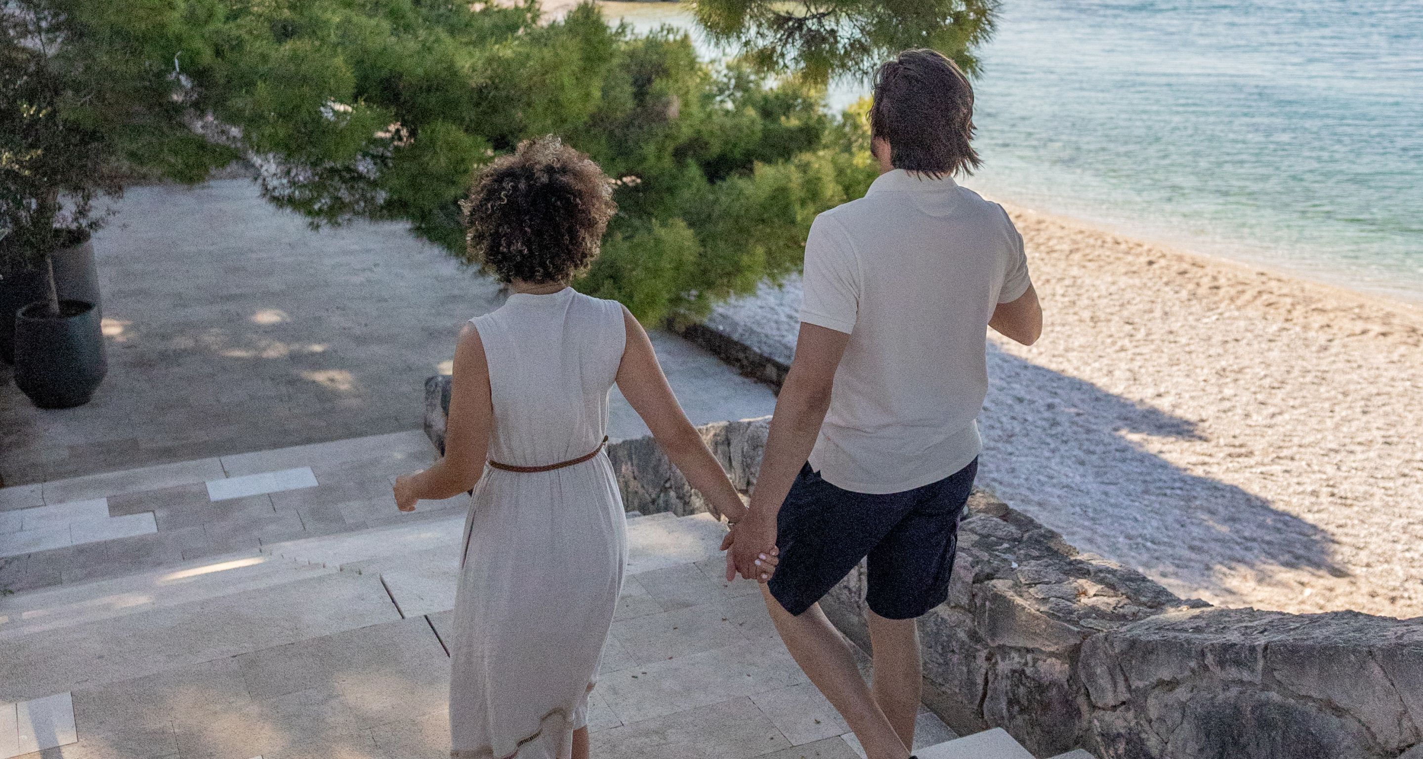 A couple walking hand in hand on a stone staircase overlooking a beach.