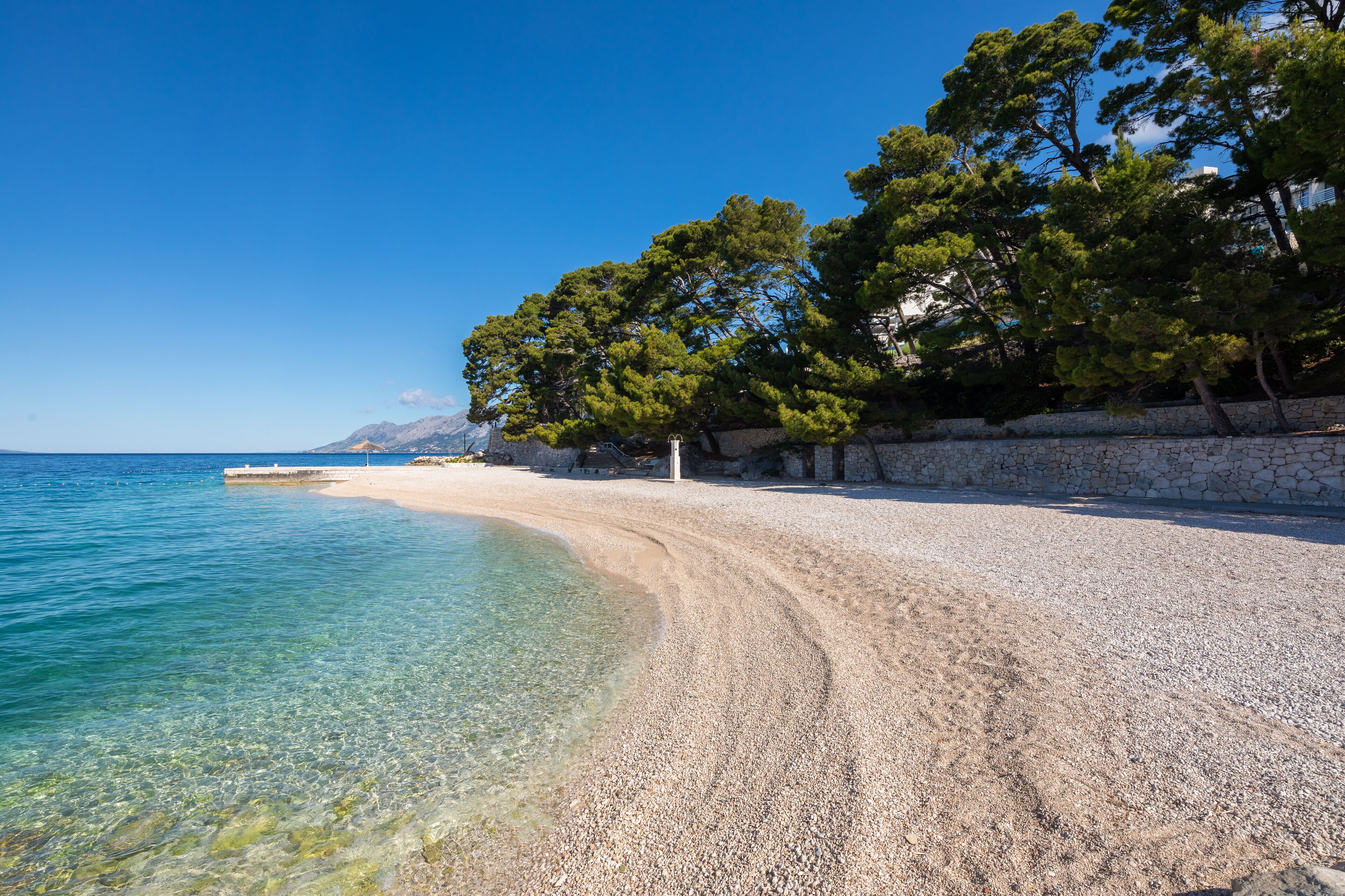 Een kiezelstrand met helder water, bomen en een stenen muur.