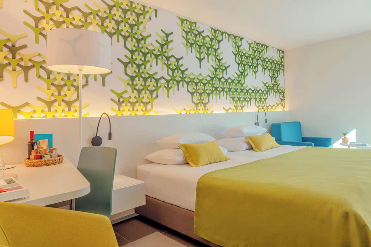 A modern hotel room with a large bed, a desk with a lamp, and a wall-mounted light fixture.
