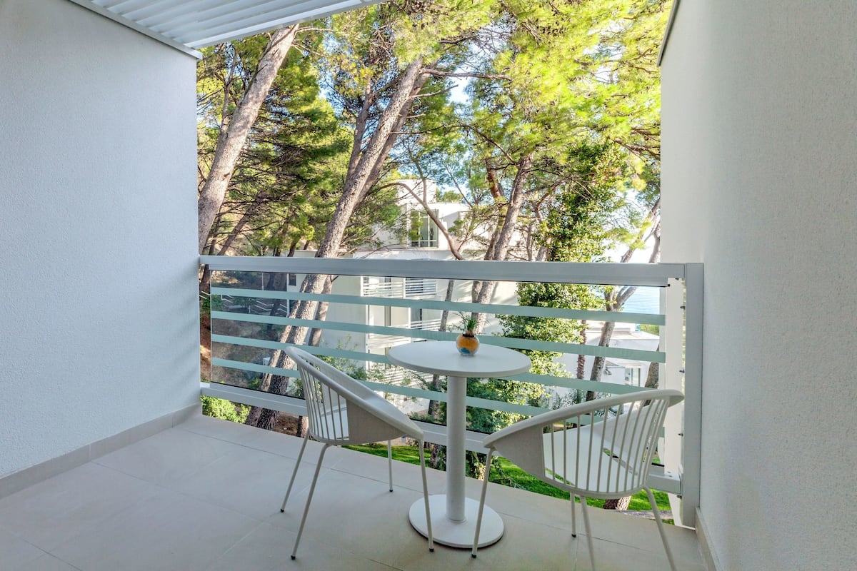 A balcony with a white table and two white chairs, a white railing, and a view of trees and a building.