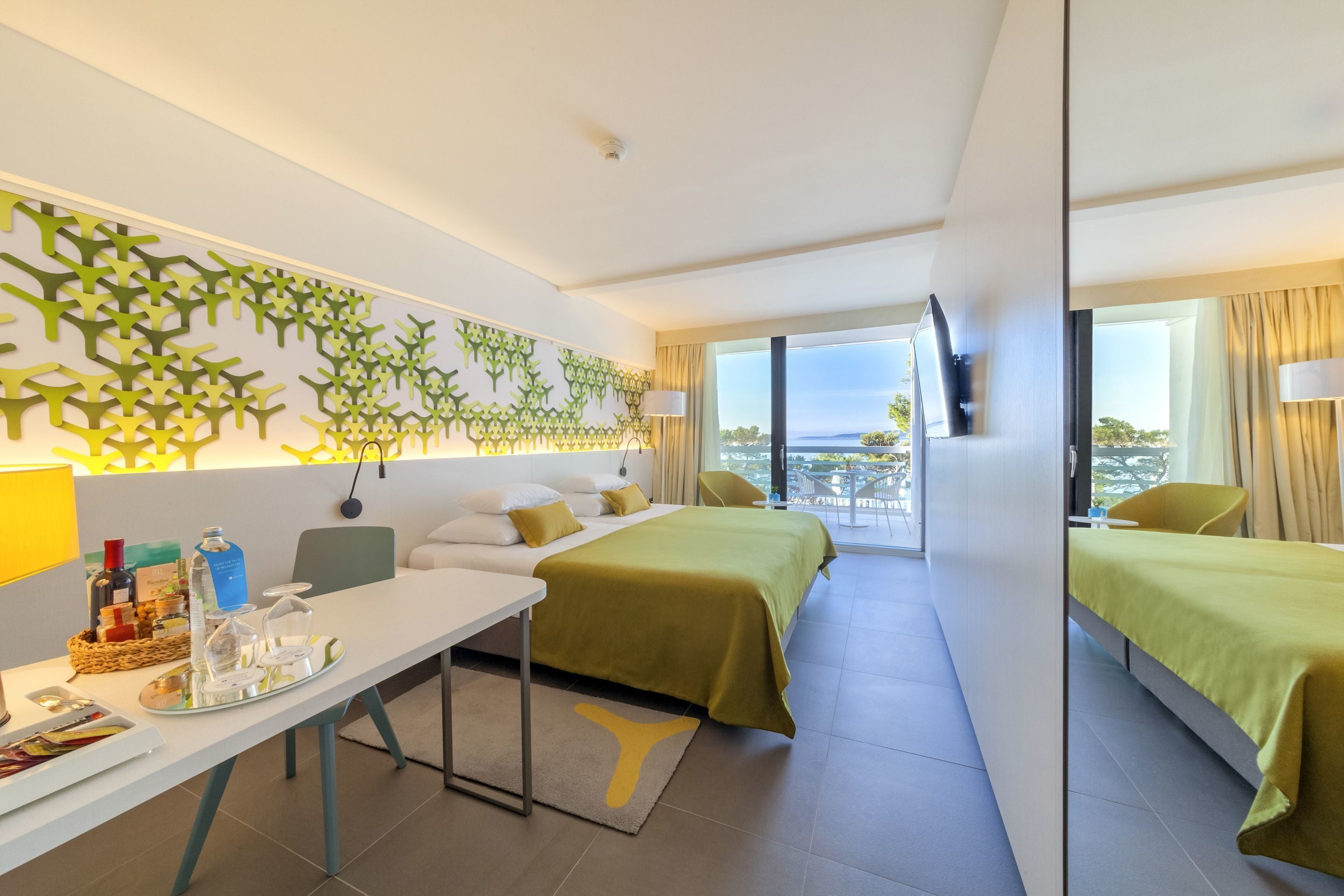 A modern hotel room with a large bed, a desk with amenities, a decorative wall, and a balcony with a view.