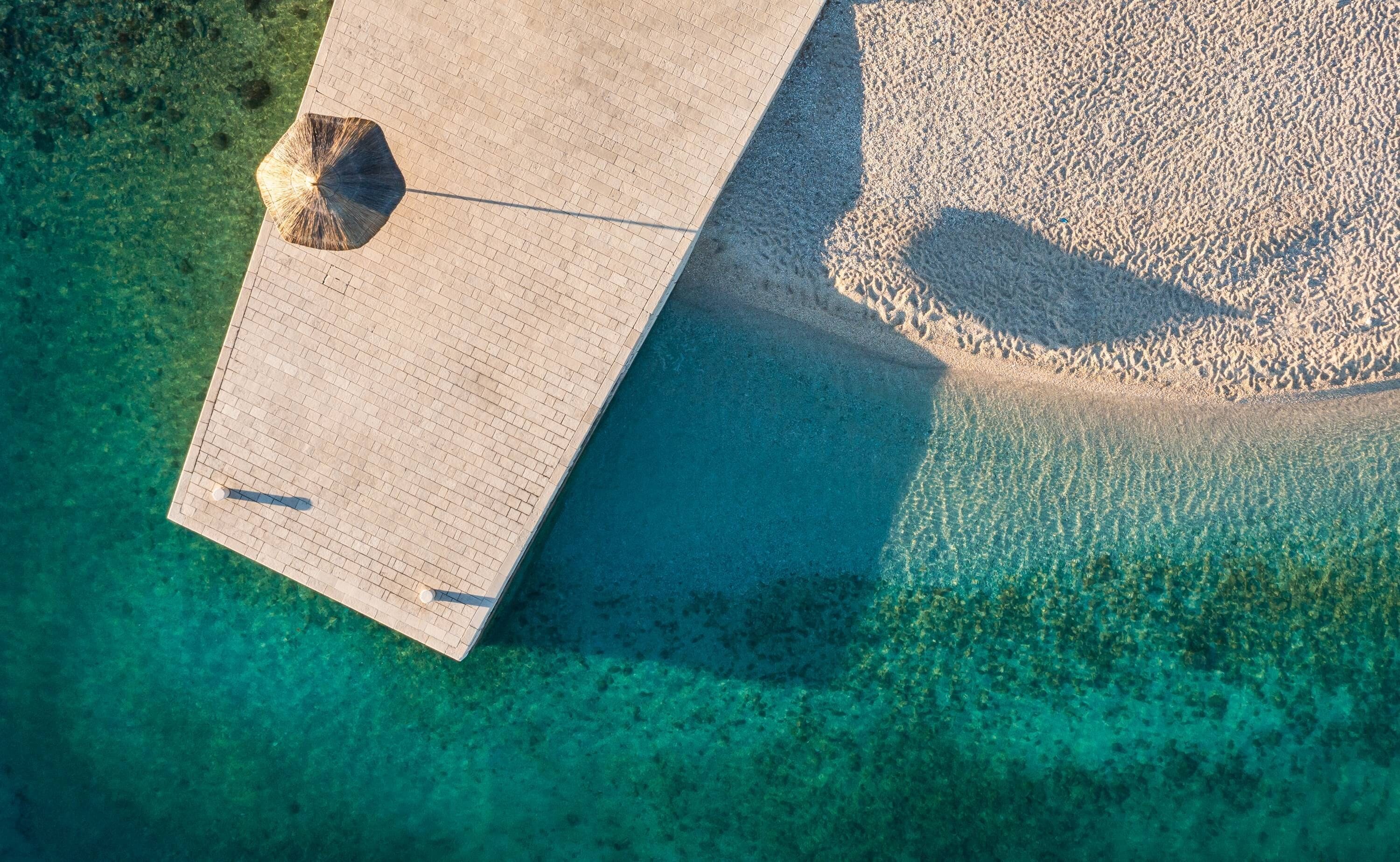 An aerial view of a beach with clear turquoise water, a paved walkway, and a large umbrella.