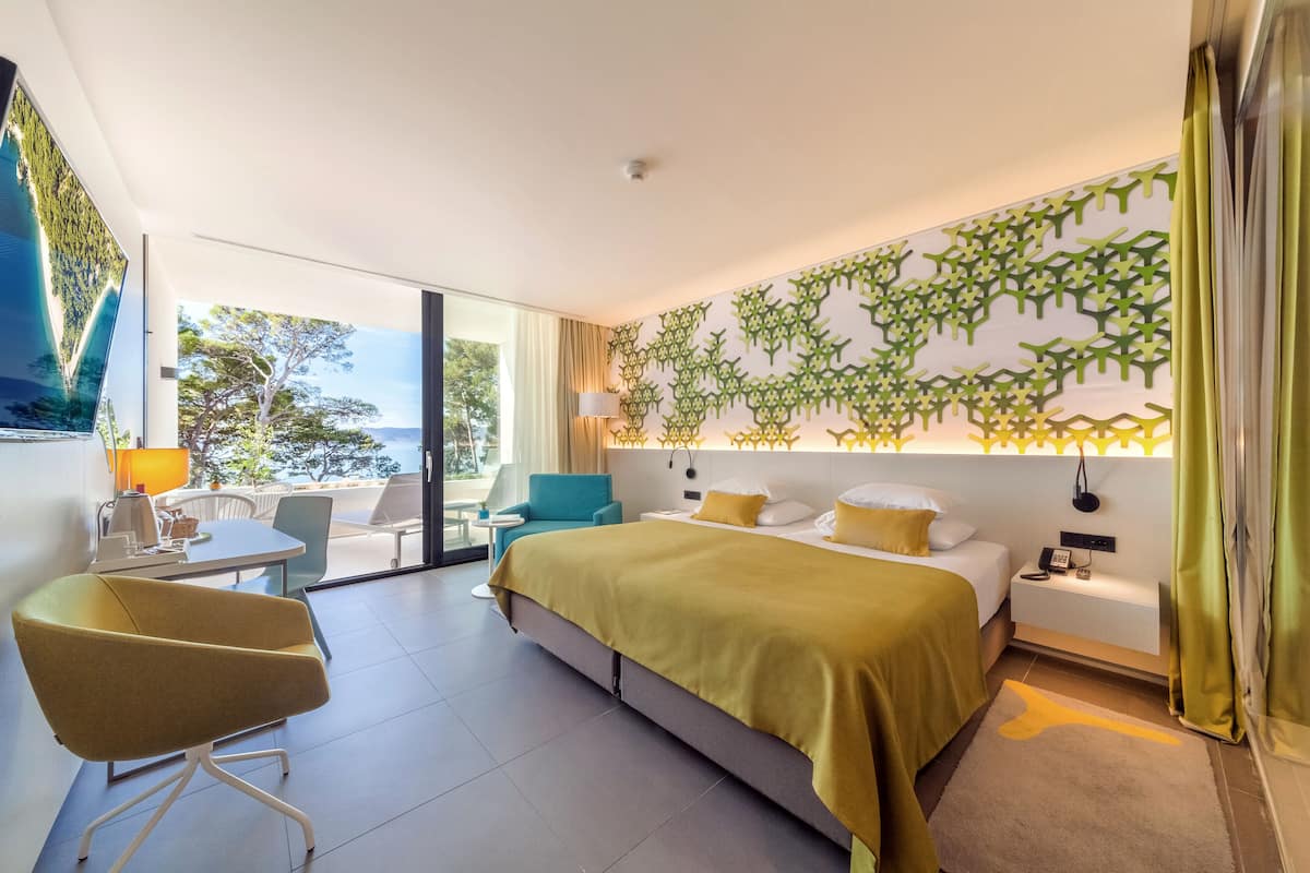 A modern hotel room with a large bed, a desk with a chair, a small table, and a view of the outdoors.