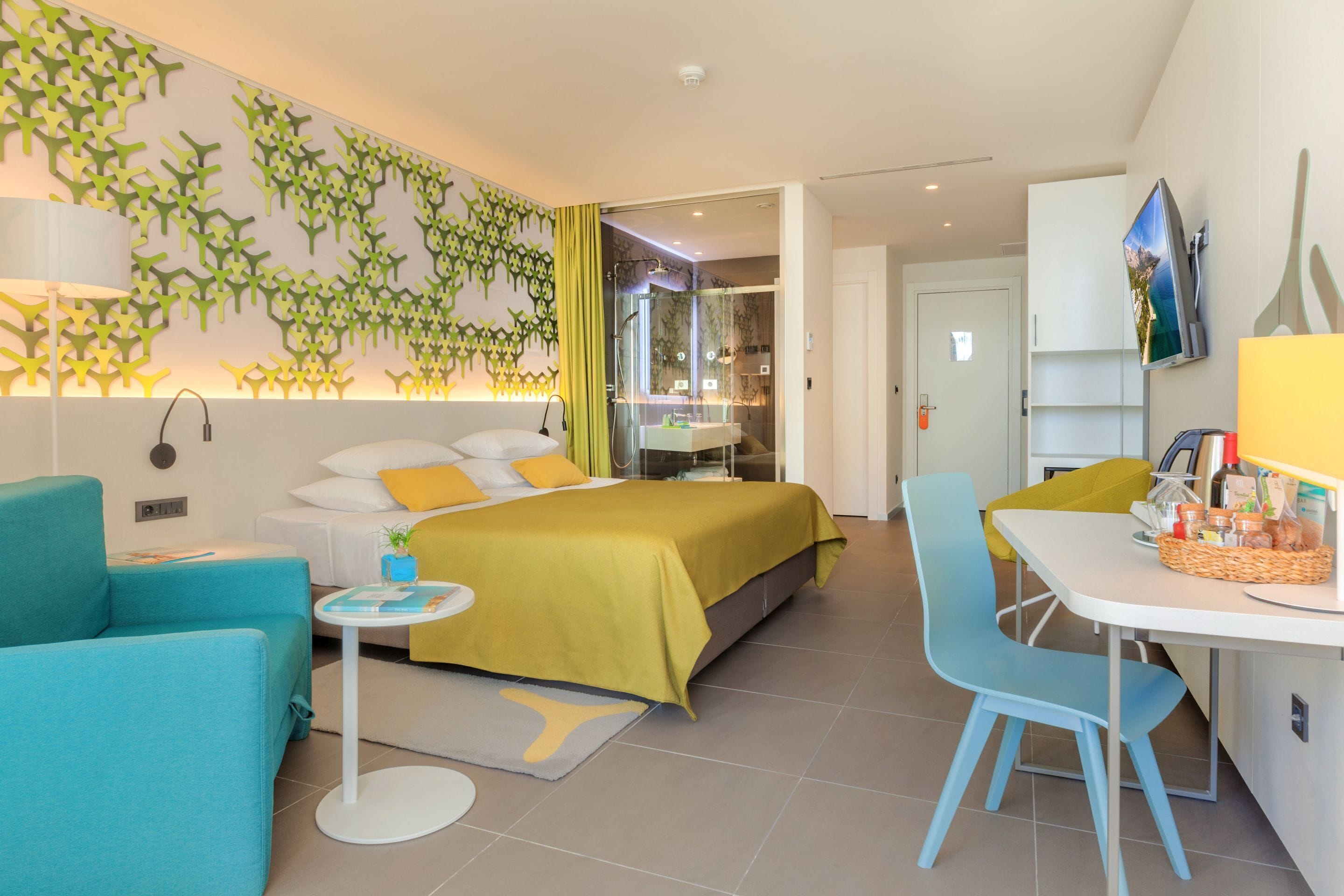 Ein modernes Hotelzimmer mit einem Bett, einem kleinen Tisch, einem Sessel, einem Fernseher und einem durch eine Glastrennwand abgetrennten Badezimmer.