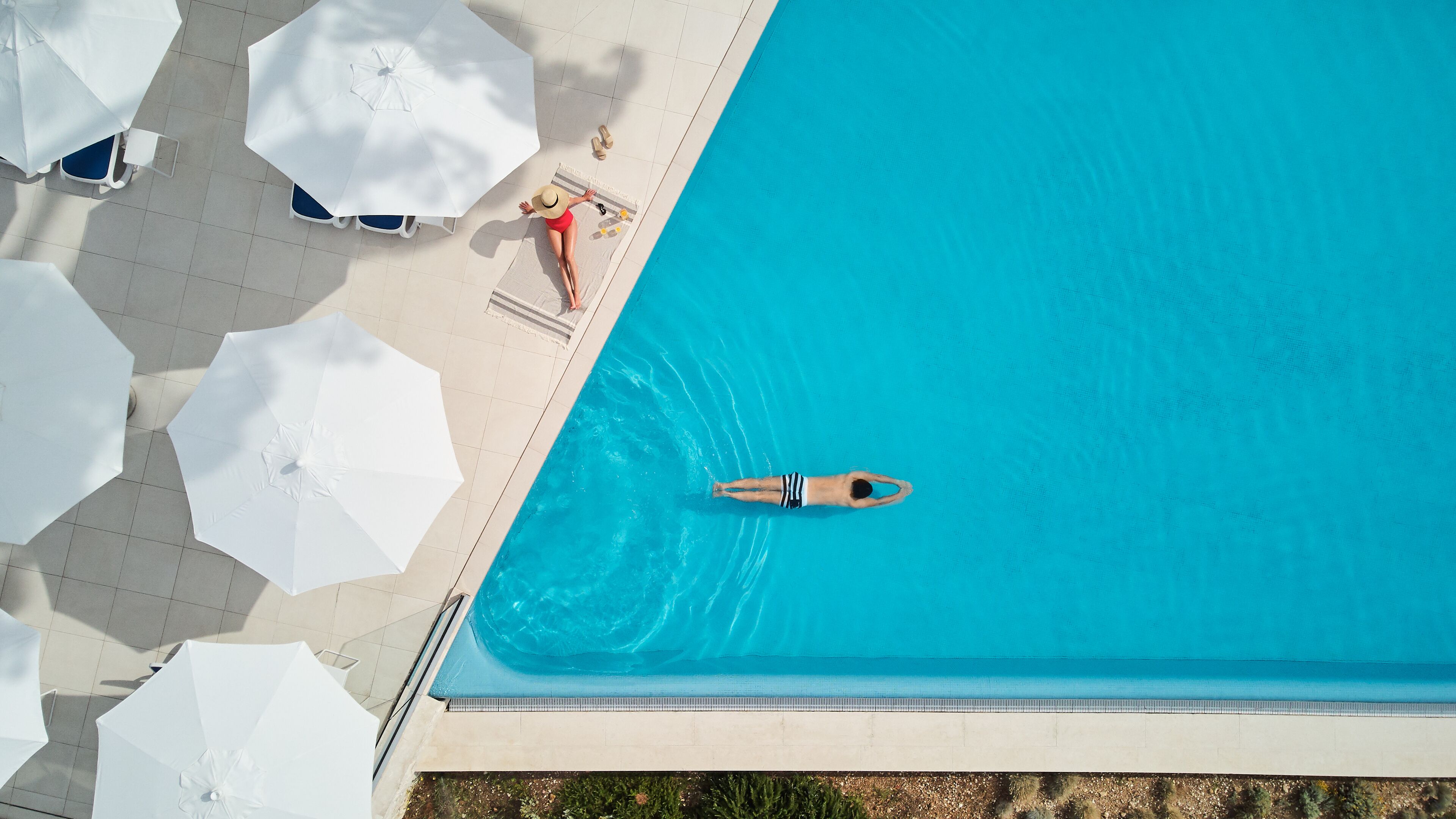 An aerial view of a swimming pool with clear blue water, surrounded by white umbrellas and lounge chairs.