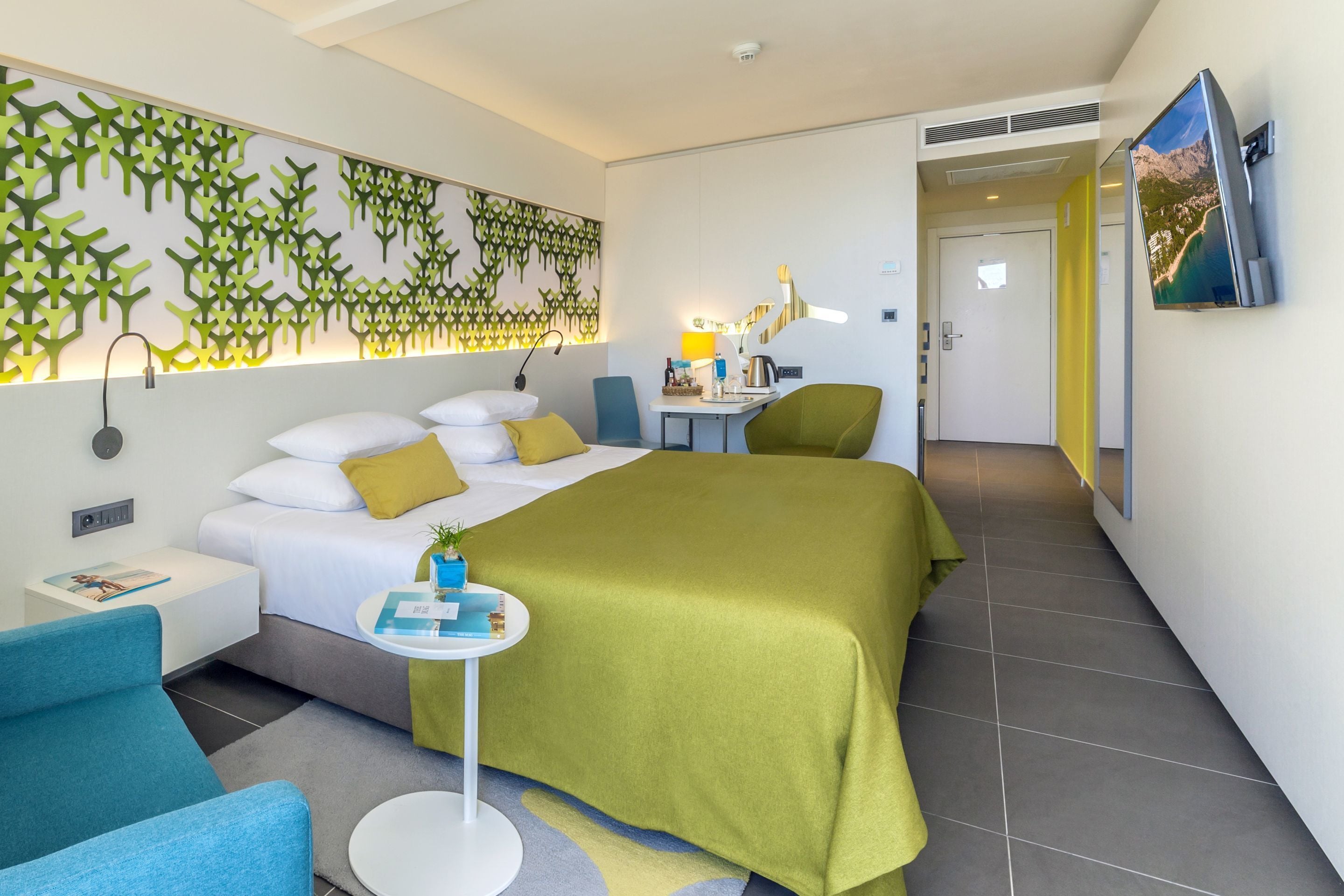 A modern hotel room with a large bed, a desk, a chair, a TV, and a decorative wall.