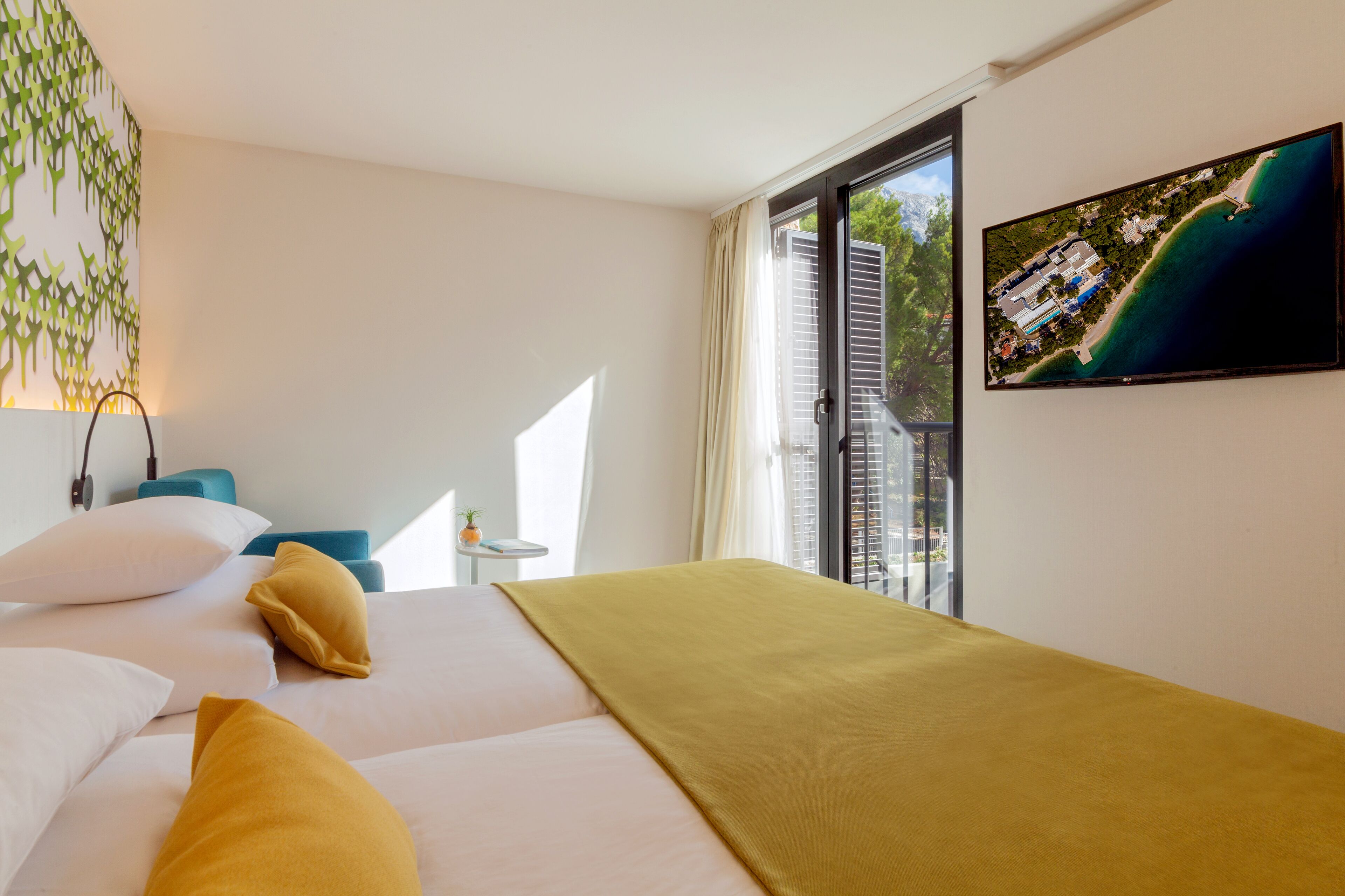 A hotel room with two beds, a television, a small table, and a window with a view of a coastal area.