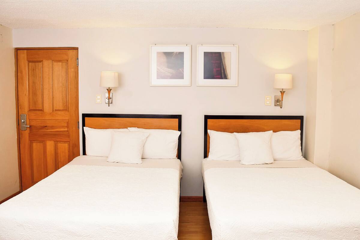 A hotel room with two single beds, wooden headboards, white bedding, two wall-mounted lamps, and a wooden door.
