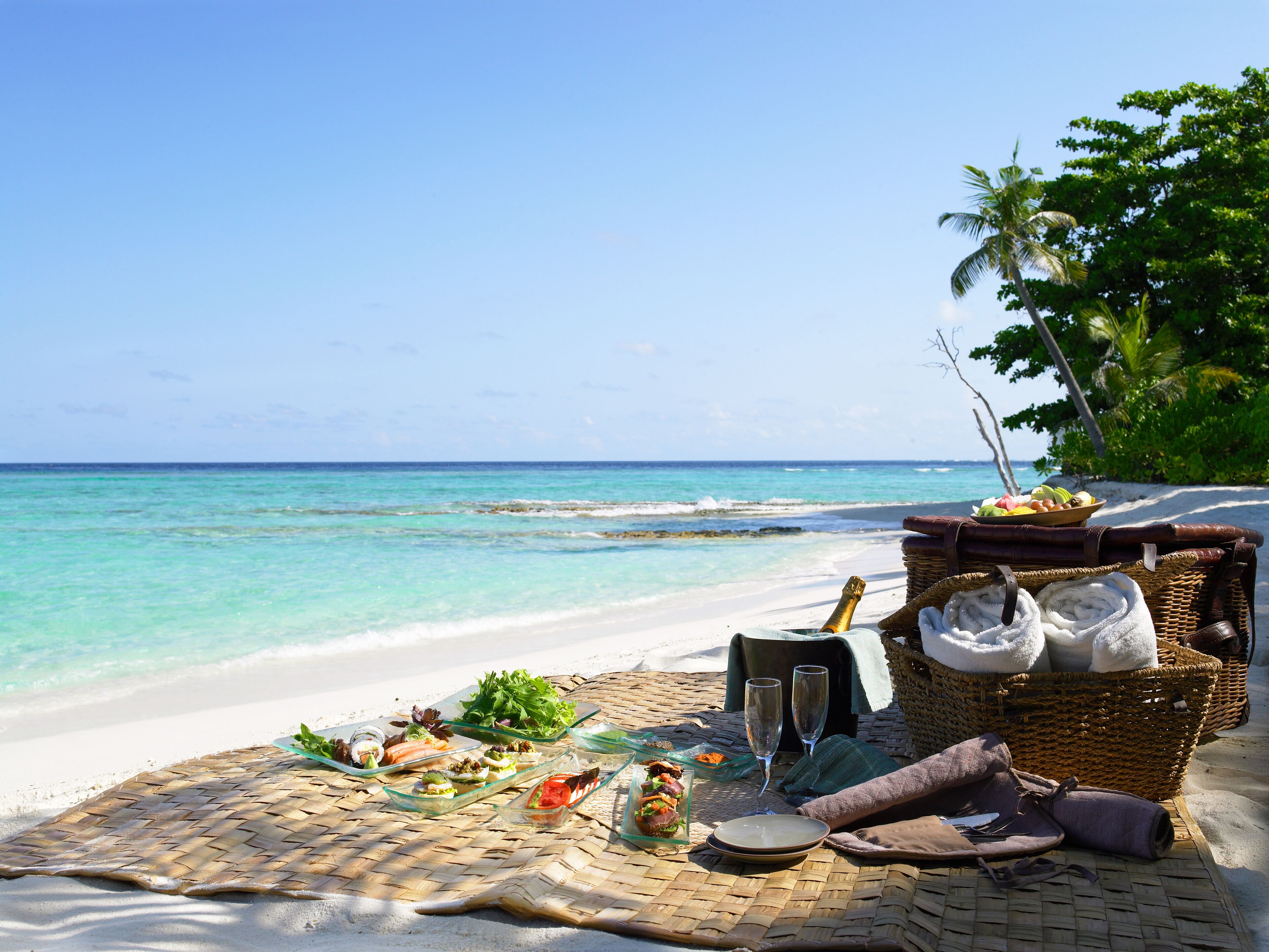 Un montaje para picnic en la playa con comida, bebidas y toallas sobre una estera tejida.