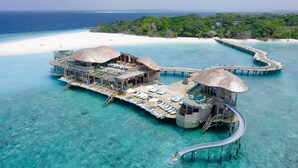 Villas sobre el agua con múltiples terrazas, agua turquesa cristalina y una playa de arena blanca al fondo.