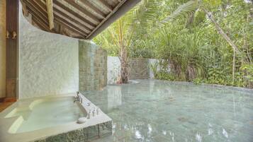 Un área de spa con suelo de baldosas de piedra, una bañera de hidromasaje y exuberante vegetación al fondo.