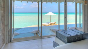 Habitación con puerta de vidrio grande que da a una piscina y vista a la playa. Hay dos sillones con almohadones, una sombrilla blanca y una maleta grande sobre la cama.
