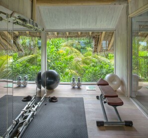A gym with exercise equipment, a bench, and a view of lush greenery through large windows.