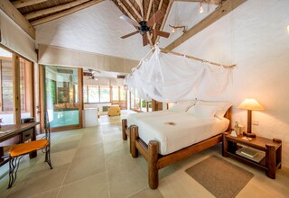 A spacious bedroom with a large bed, wooden headboard, and a canopy. There is a bedside table with a lamp, a chair, and a small table with a lamp. The room has a wooden ceiling and large windows.