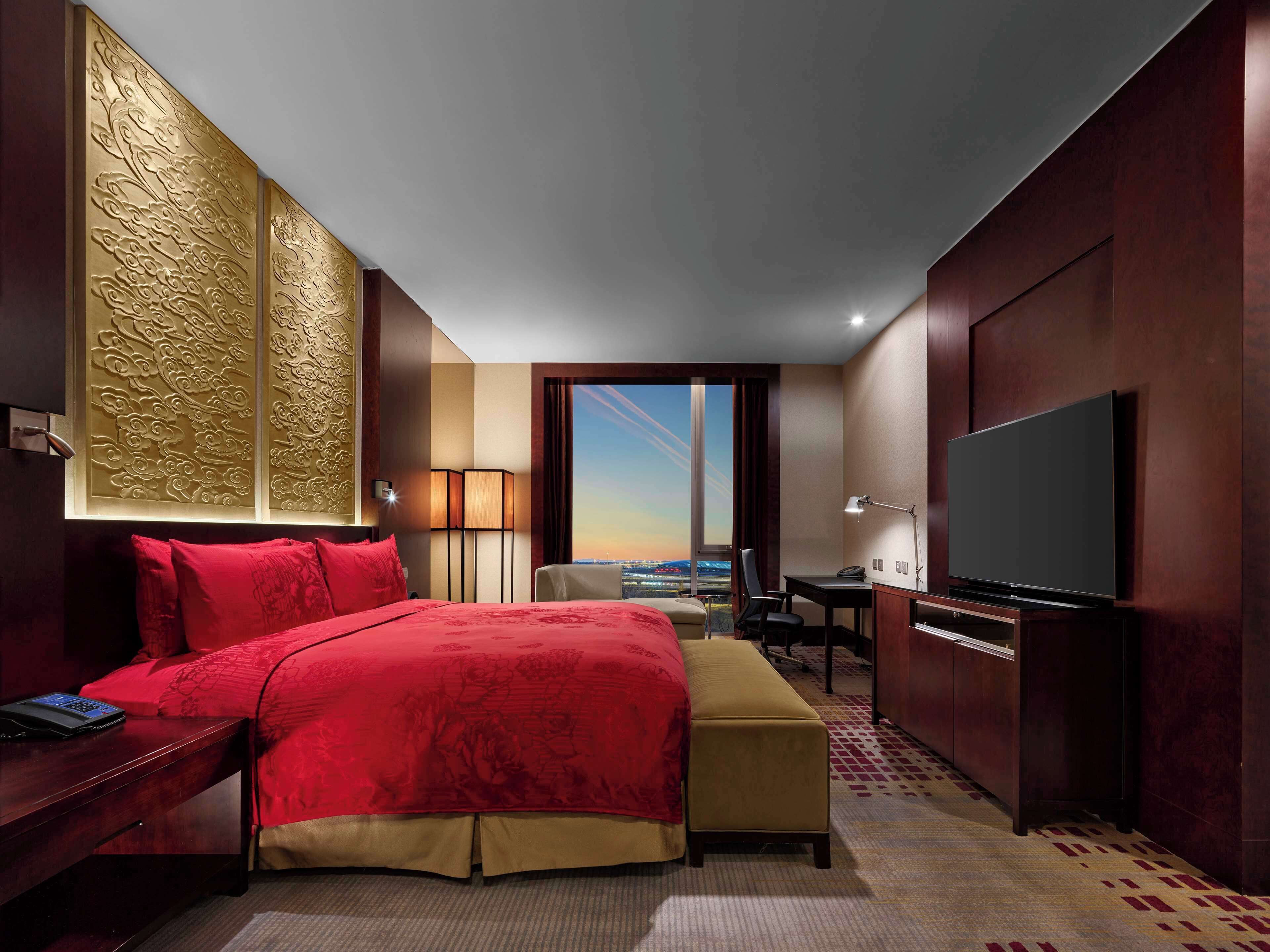 A hotel room with a large bed, a desk, a chair, a TV, and a view of the cityscape.