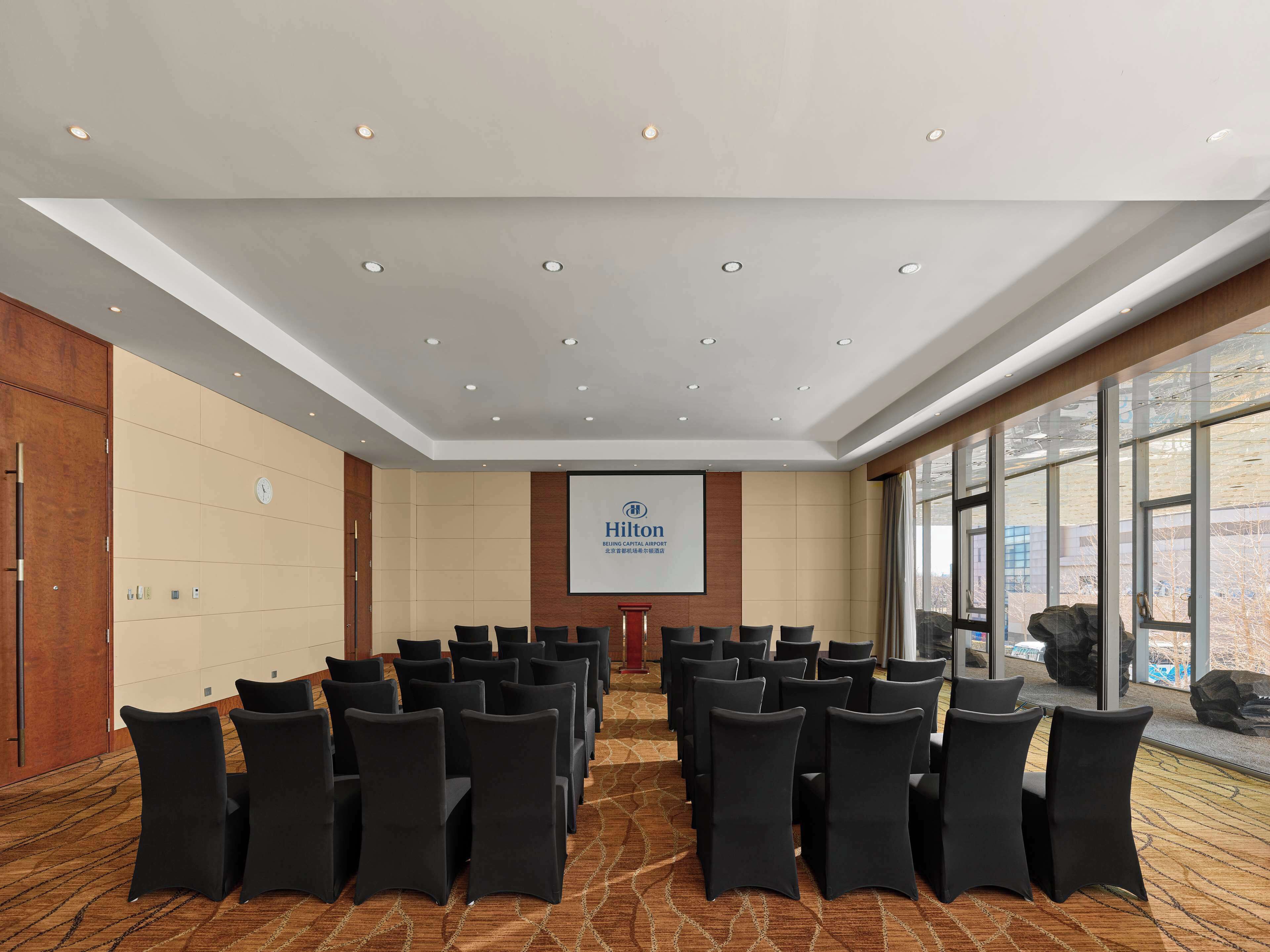A conference room at the Beijing Capital International Airport Hilton with rows of chairs, a podium, and a large screen displaying the Hilton logo.
