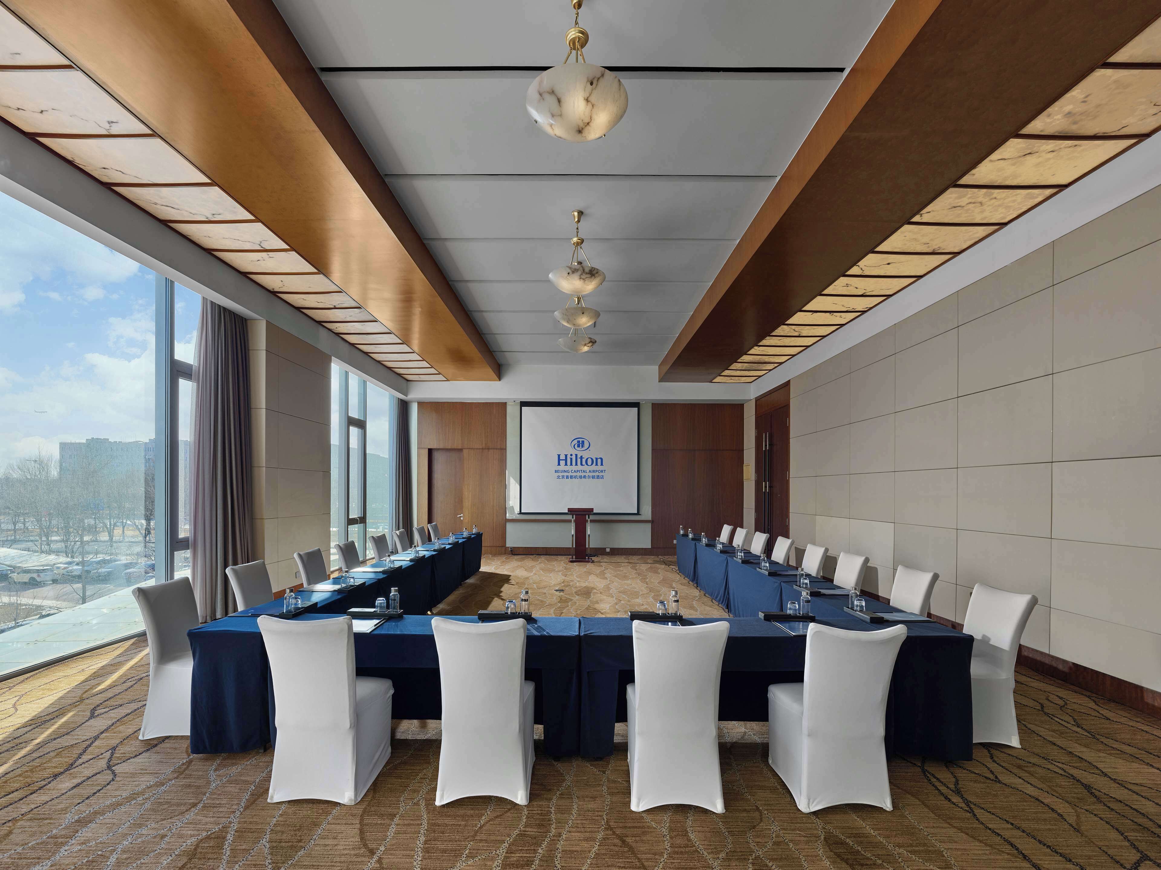 Una sala conferenze con un lungo tavolo, sedie bianche e uno schermo proiettore che mostra "Hilton".