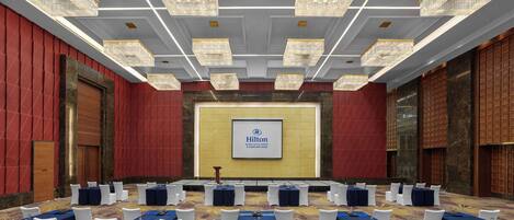 A conference room with a large screen displaying the Hilton logo, multiple tables with white chairs, and chandeliers.