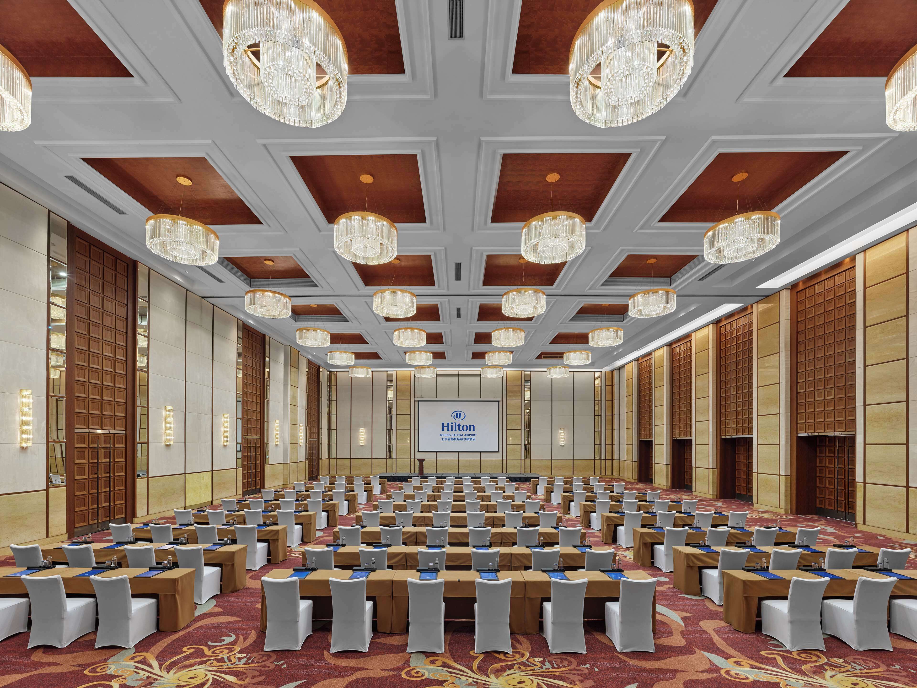 A large conference room with rows of tables, white chairs, and chandeliers.