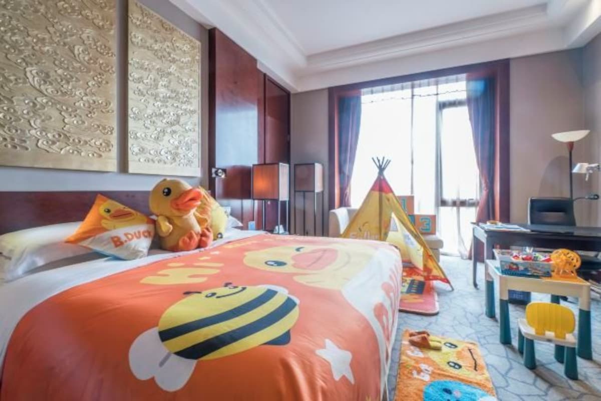 A hotel room with a large bed featuring a duck-themed bedspread, a small play tent, and a toy table with a toy lion.