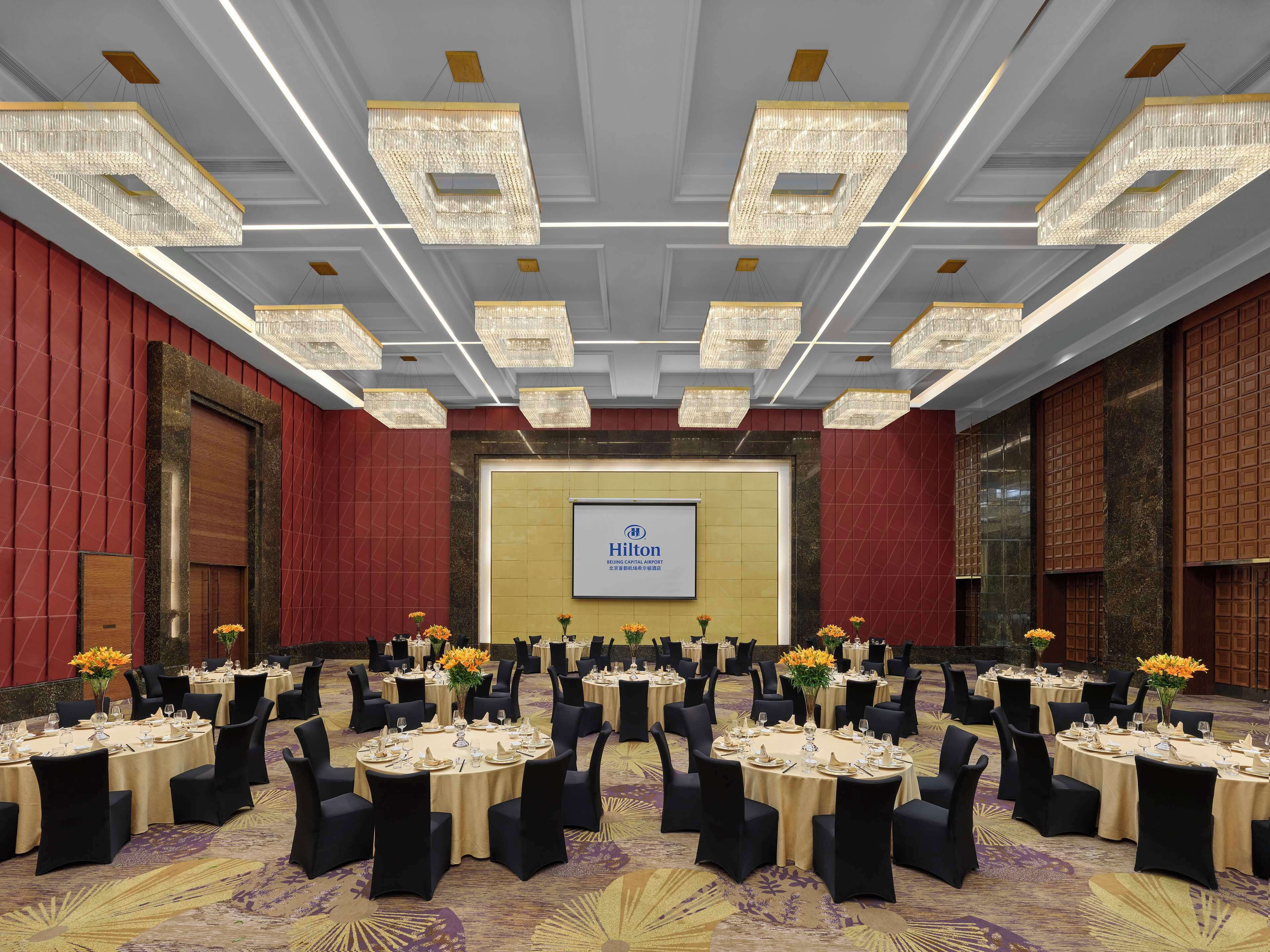 Een ruime congreszaal met ronde tafels, zwarte stoelen en kroonluchters. Een groot scherm toont het Hilton logo.