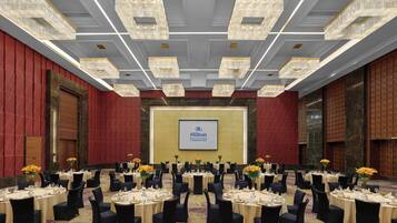 Un salón de conferencias amplio con mesas redondas, sillas negras y candelabros. Una pantalla grande muestra el logotipo de Hilton.